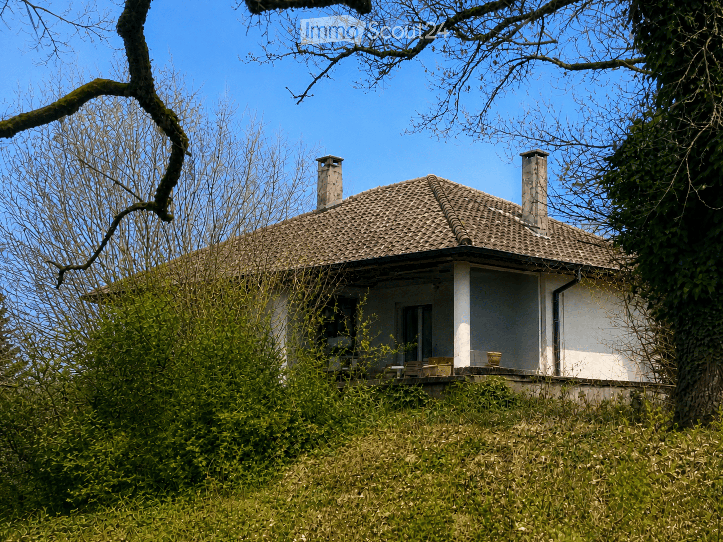 A small, single-story house with a tiled roof and two chimneys, surrounded by trees and greenery.