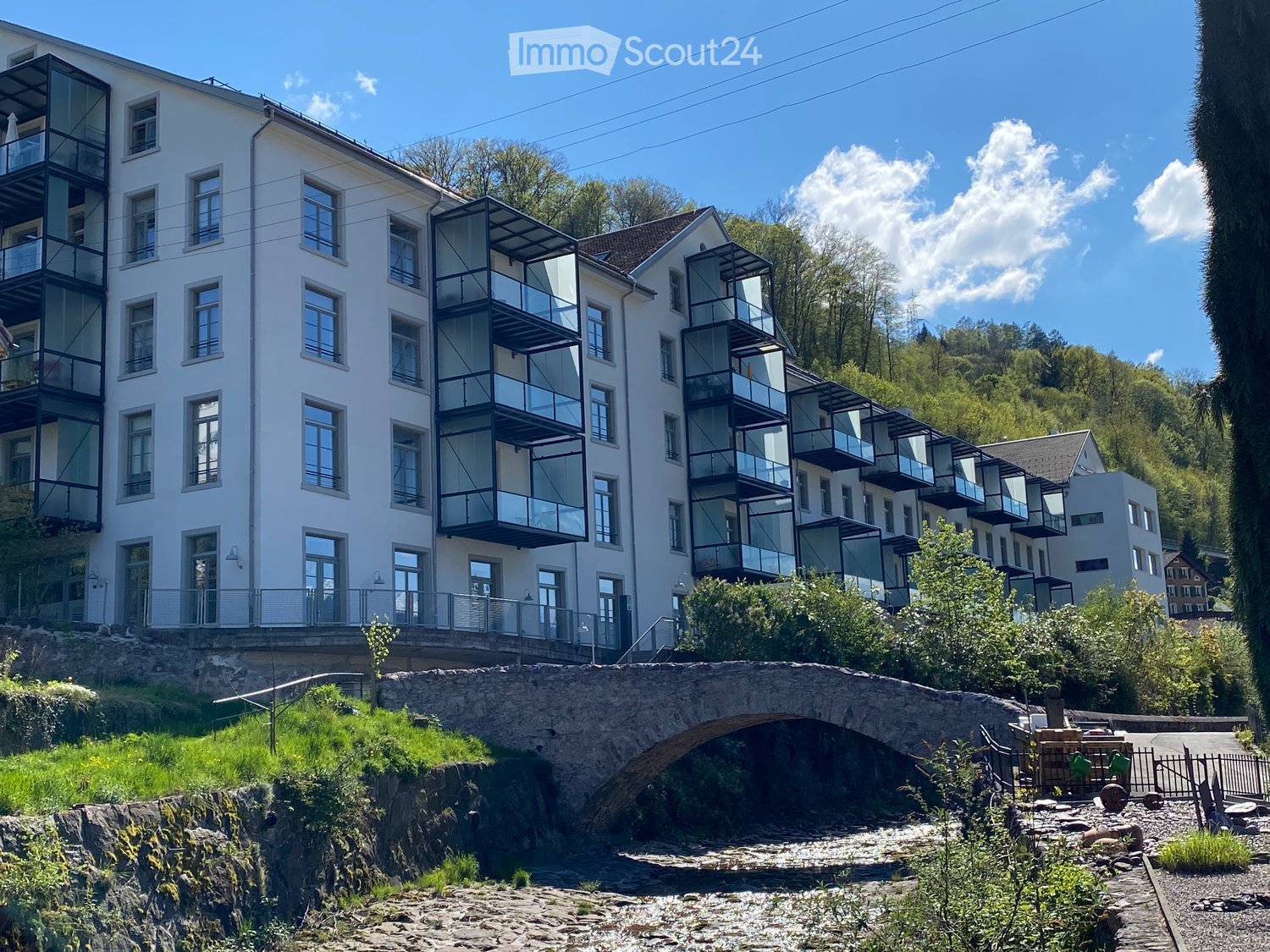 Modern multi-story building with white facade, large windows, and multiple balconies overlooking a river and stone bridge.