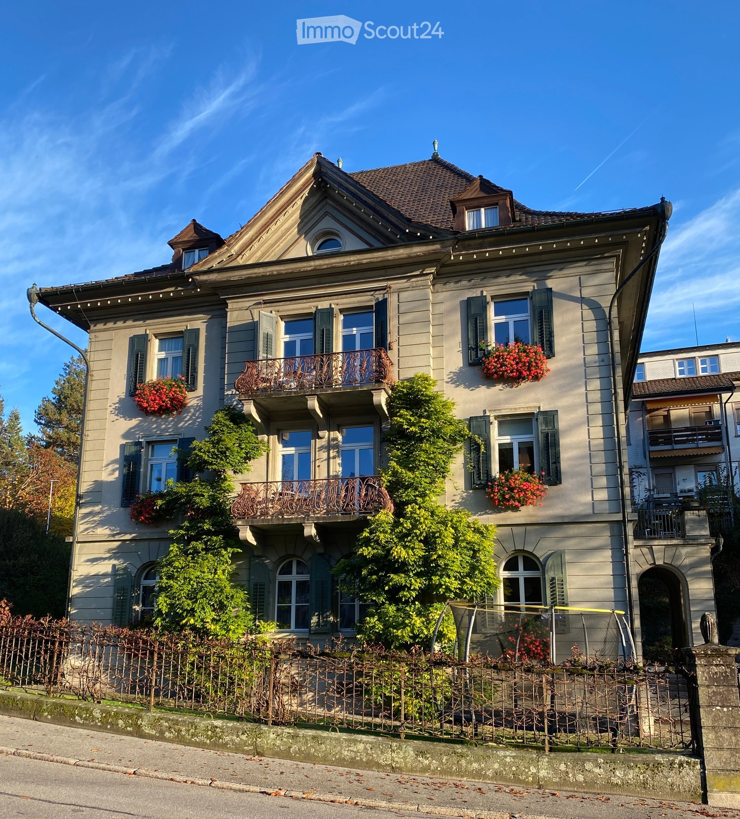Three-story residential house with beige facade, multiple windows, balconies, and decorative railings. Greenery and flower boxes adorn the building.
