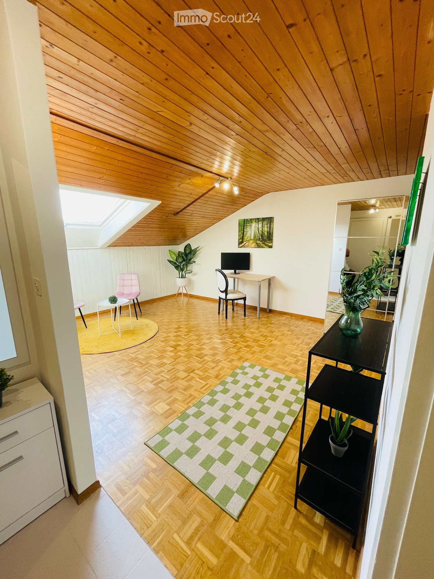 Wooden floor, wooden ceiling, white walls, desk with computer monitor, pink chair, round table, rug, potted plants, mirror, white cabinet with drawers