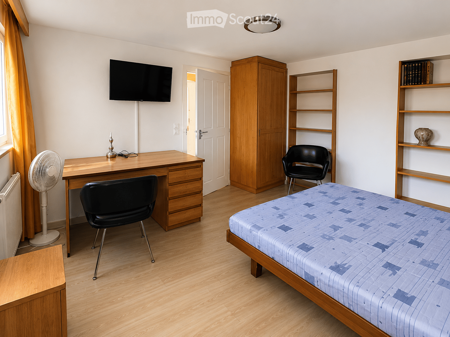 Well-lit bedroom with a single bed, wooden desk, two chairs, a fan, a cabinet, shelves, and a television mounted on the wall.