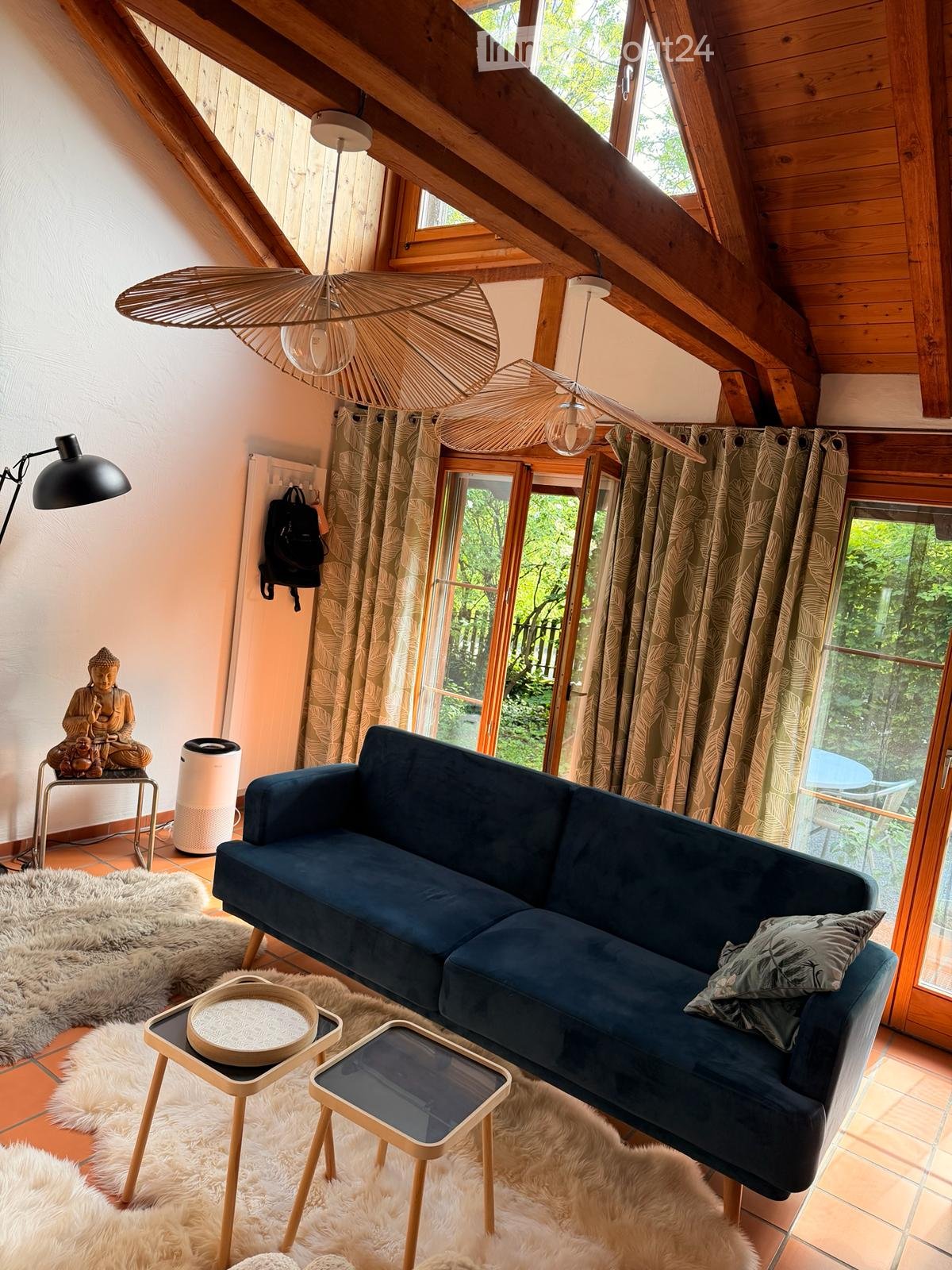 A cozy living room with a dark blue sofa, wooden stools, a round wooden table, a large window with curtains, and a decorative rug on the floor.