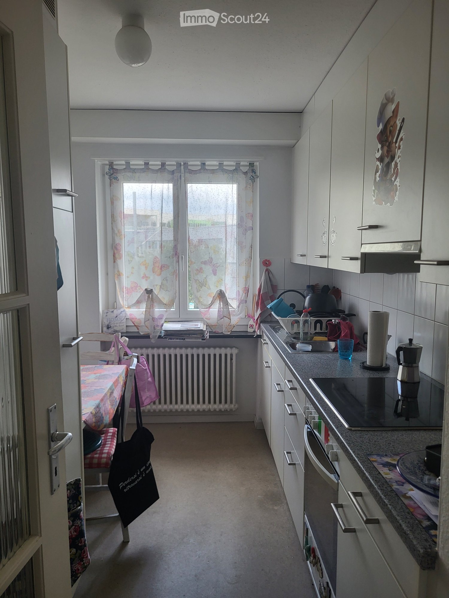 A small kitchen with white cabinets, a stove, sink, and a table with a colorful tablecloth. The window has butterfly-patterned curtains, and there are various kitchen items on the counter.
