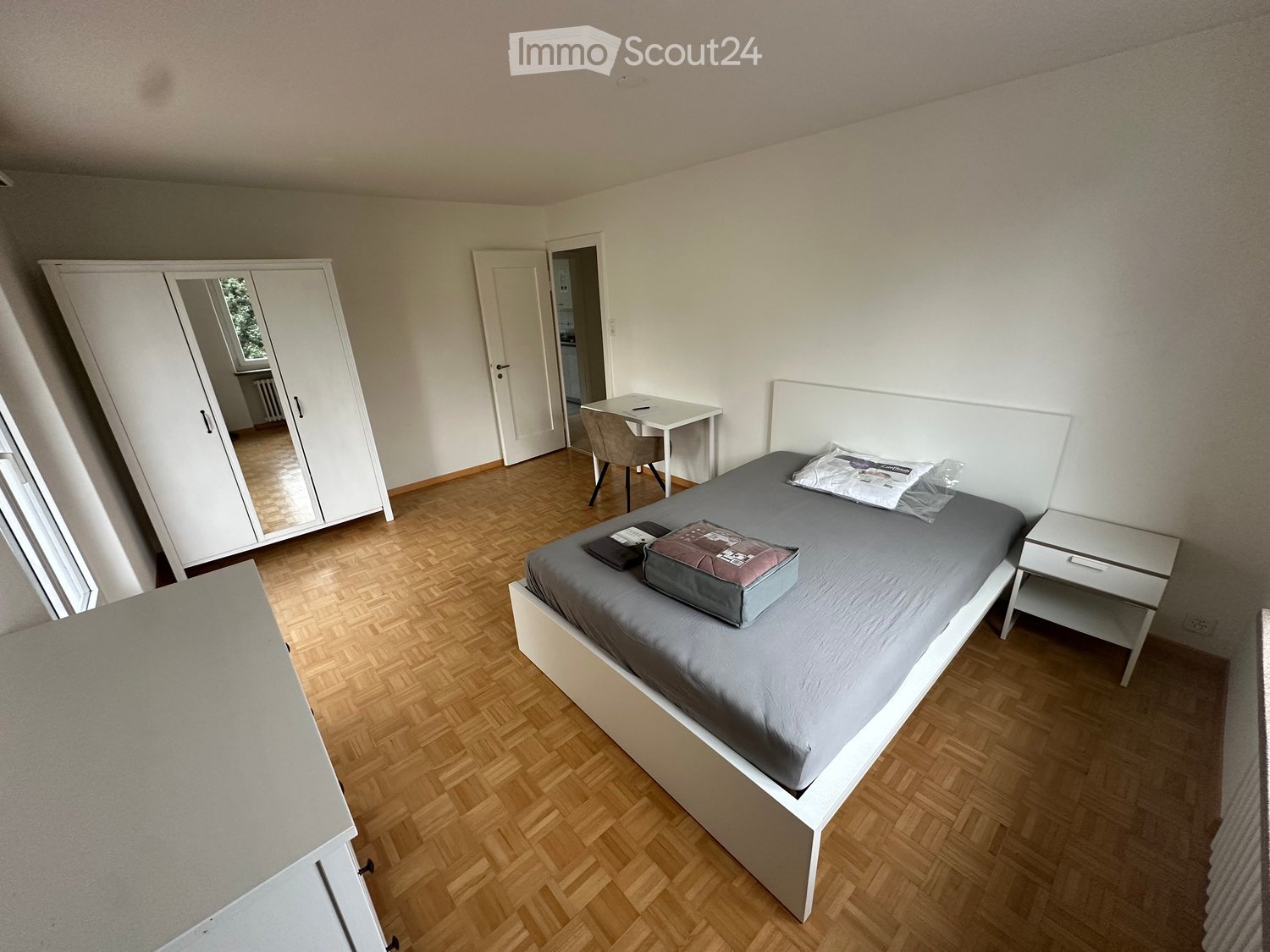 Modern bedroom with a grey duvet, wooden floor, white wardrobe, desk, and nightstand.