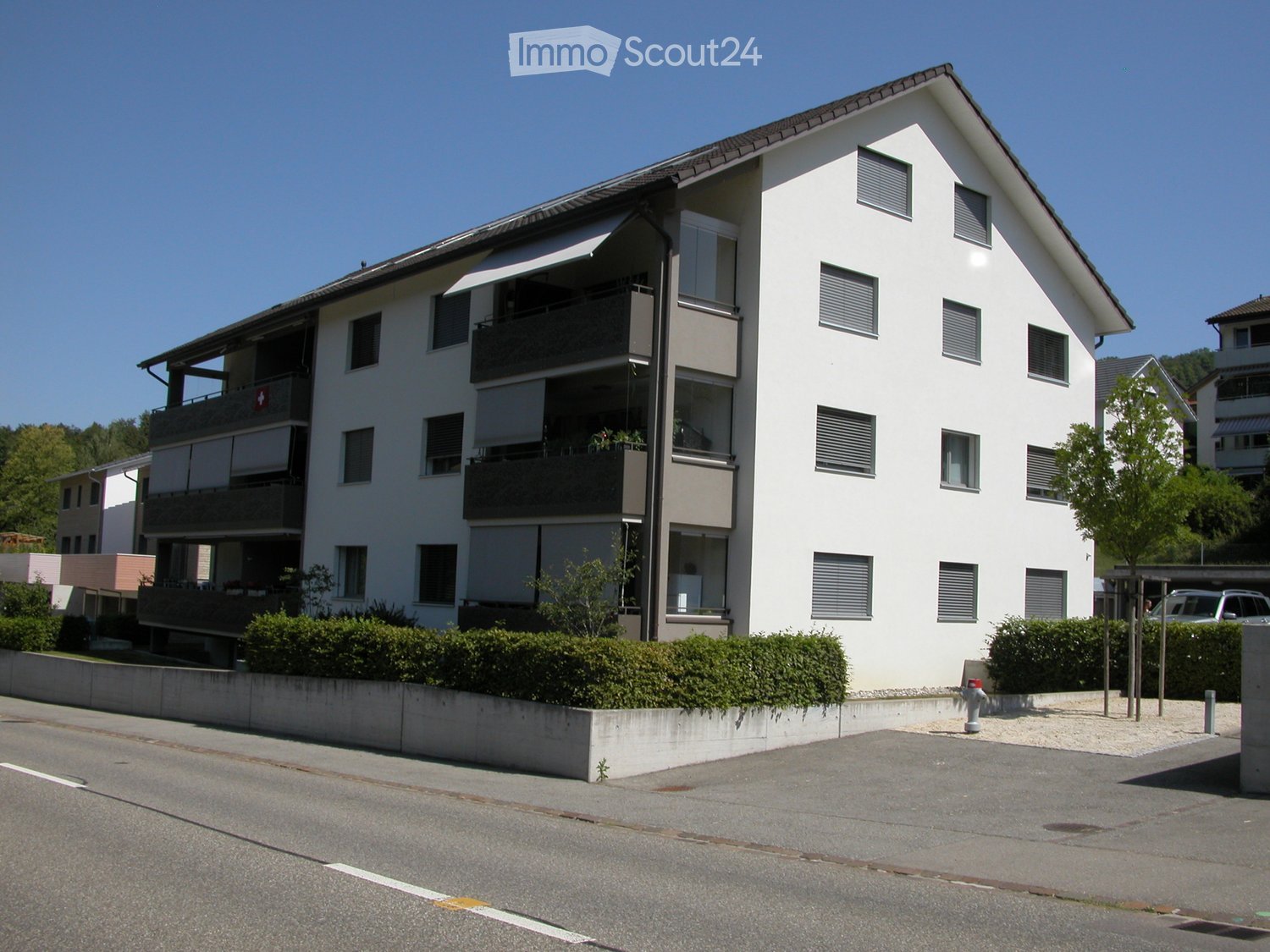 Modern white multi-story residential building with balconies, large windows, and greenery in front.