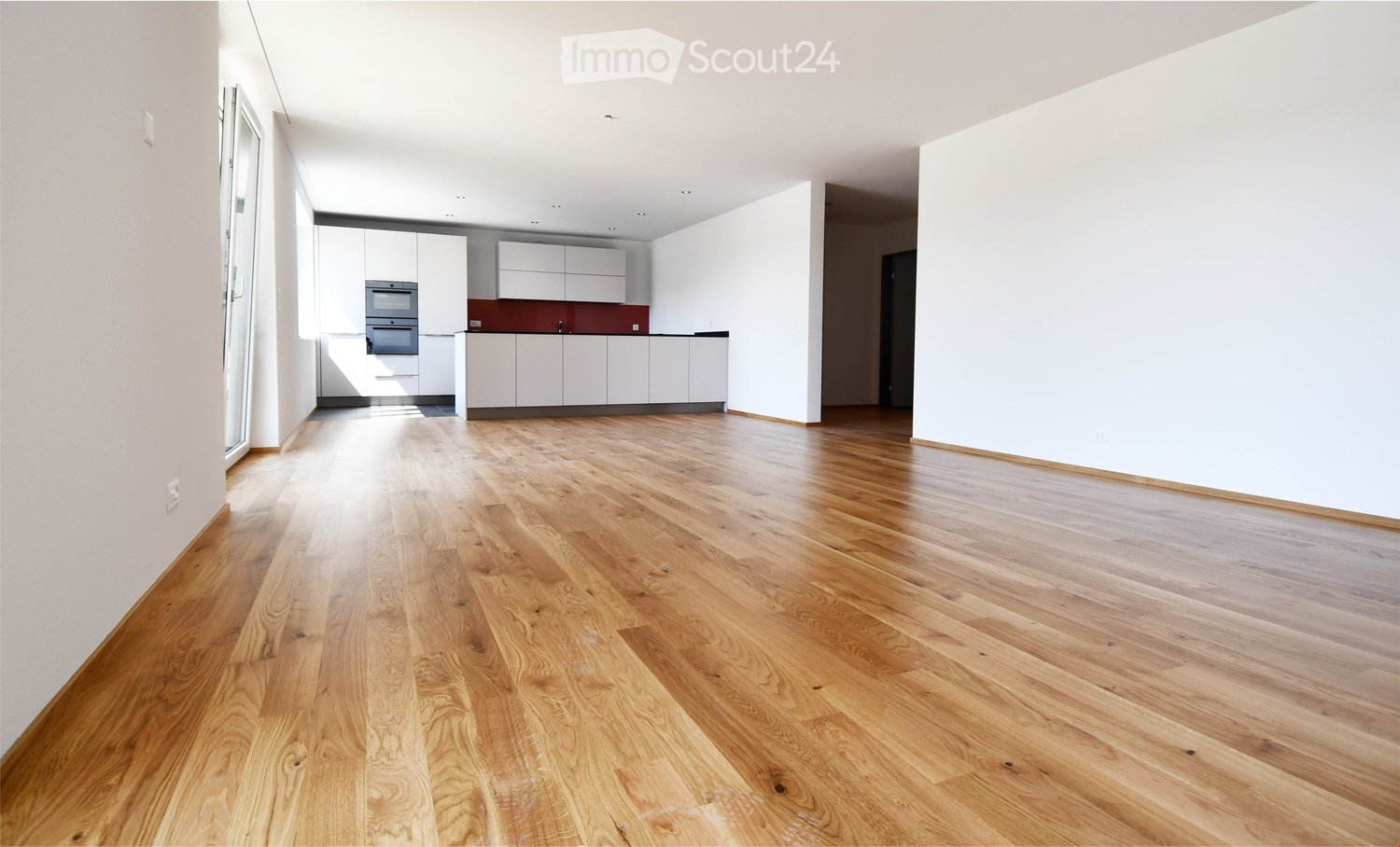 Empty room with light wooden flooring, white walls, and a modern kitchen area visible in the background.