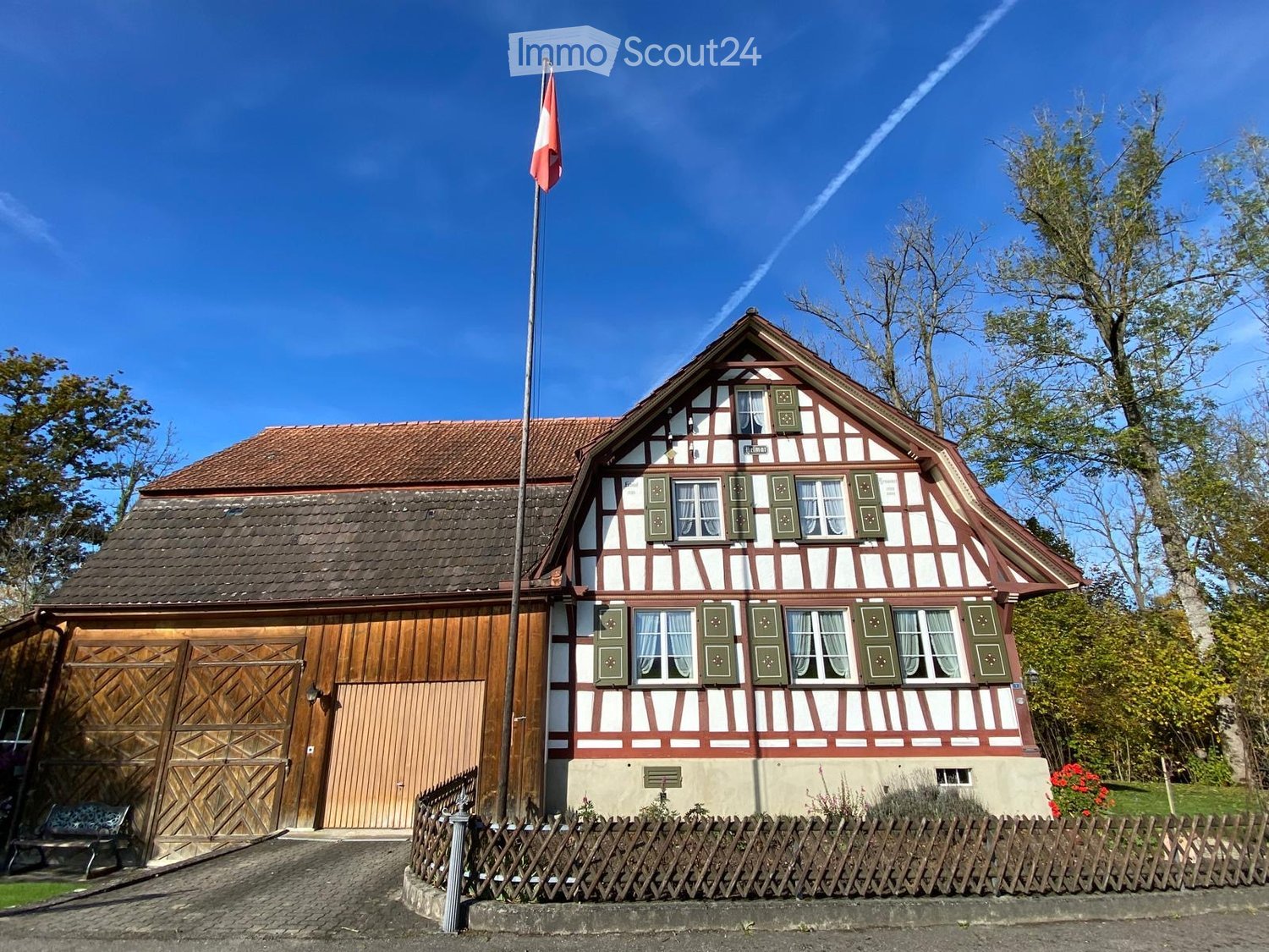 Traditional half-timbered house with red-brown roof, white walls, green shutters, and a wooden garage. A flagpole with a flag is in front, and a wooden fence encloses a small garden area.