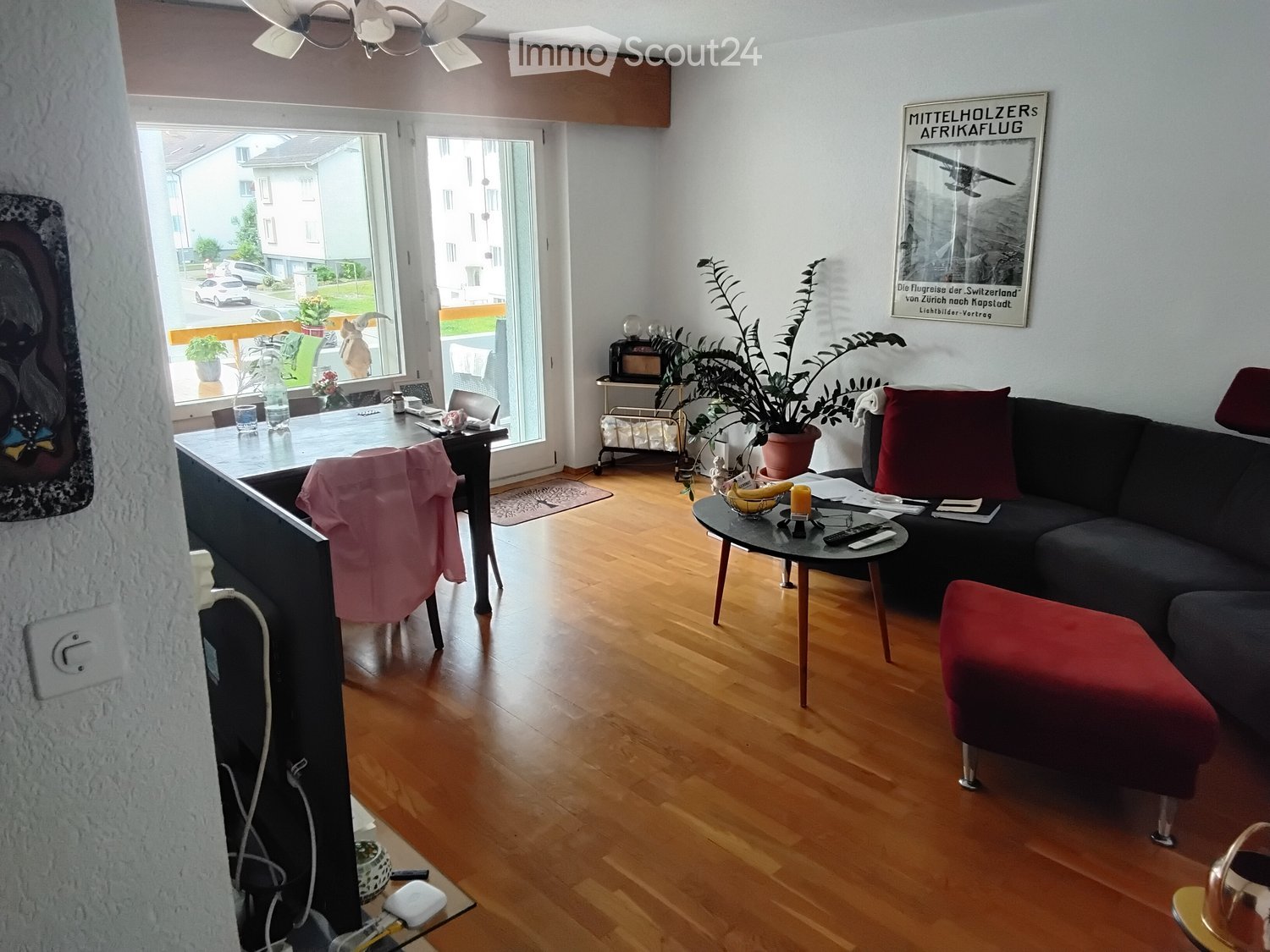 Modern living room with hardwood flooring, large windows, a dark grey sofa with red cushions, a red ottoman, a coffee table, a glass side table, a potted plant, and a framed picture on the wall.