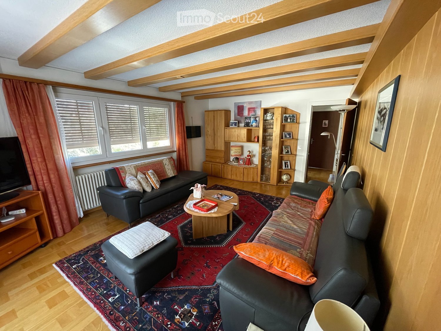 Cozy living room with wooden beams, hardwood flooring, and large windows. Features a black leather sofa, coffee table, ottoman, and a cabinet with shelves.