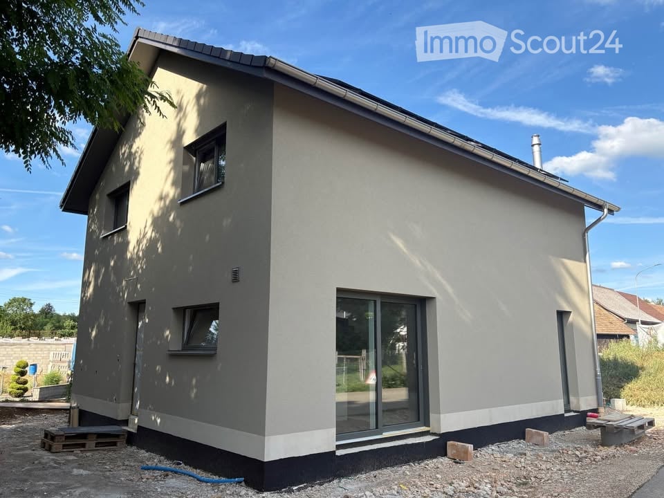Single-story house with modern design, grey and beige exterior, large glass sliding door, small windows, raised ground floor, and construction materials visible around the base.