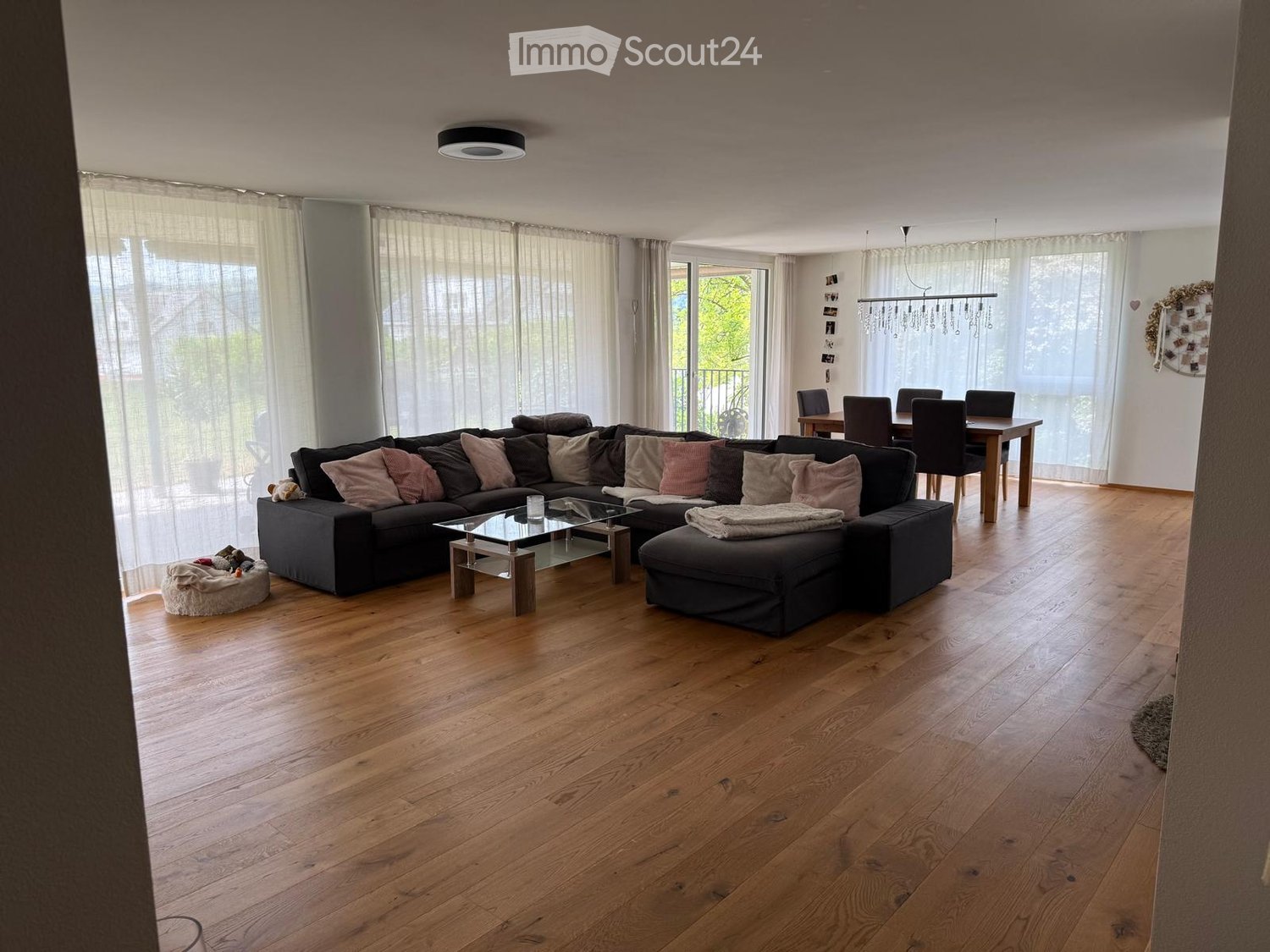 Open-plan living and dining area with wooden flooring, large windows, a sectional sofa, coffee table, dining table, and chairs.