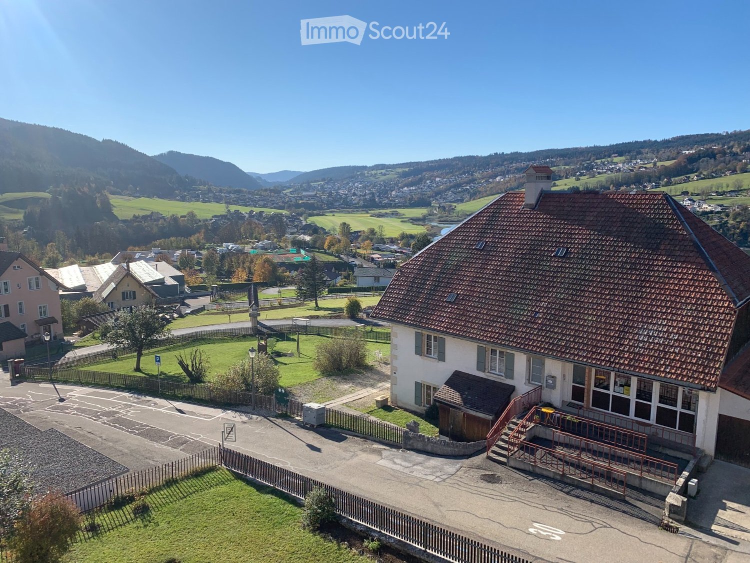 Aerial view of a two-story house with a red-tiled roof, surrounded by a fenced yard and a parking area, with a scenic valley and town in the background.