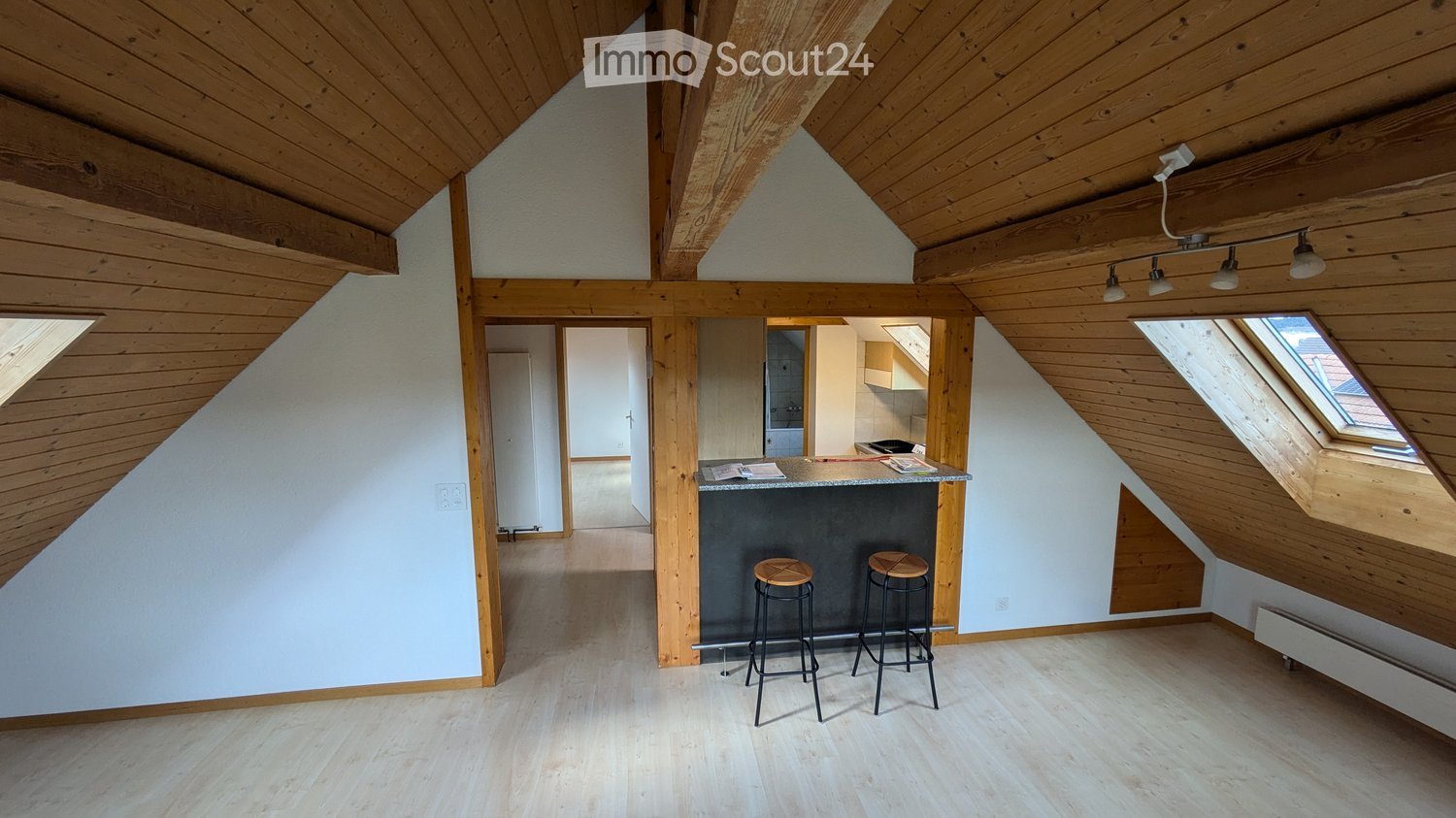 Loft-style room with wooden beams, exposed ceiling, bar counter, two stools, and modern lighting.