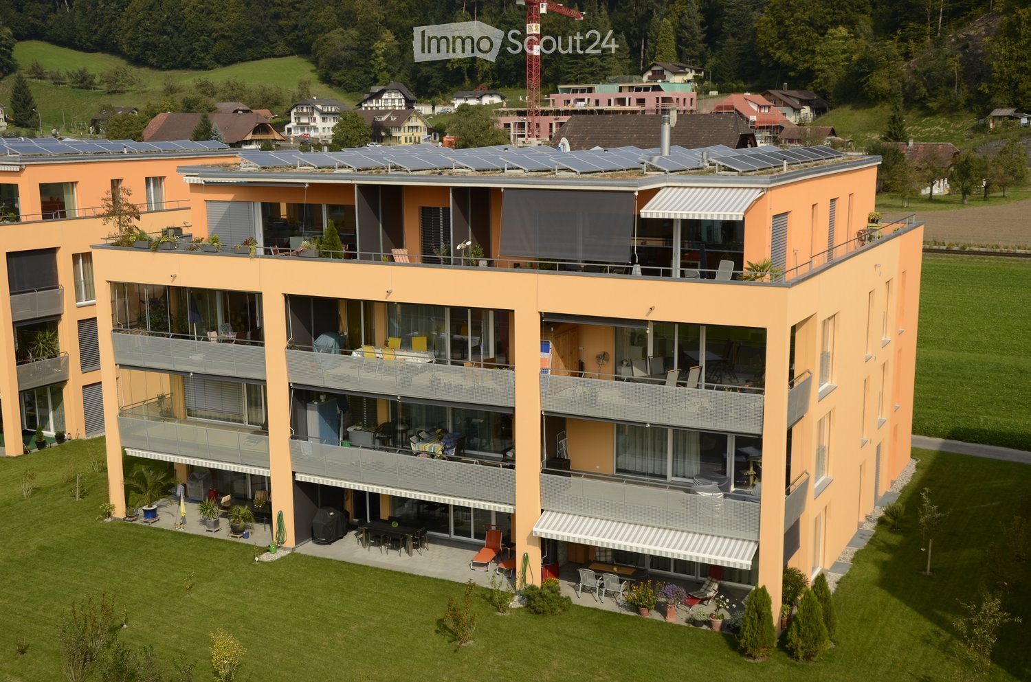 A modern, multi-story building with orange exterior walls, large windows, balconies, and solar panels on the roof. The building is surrounded by a well-maintained lawn and garden area with outdoor furniture and potted plants.