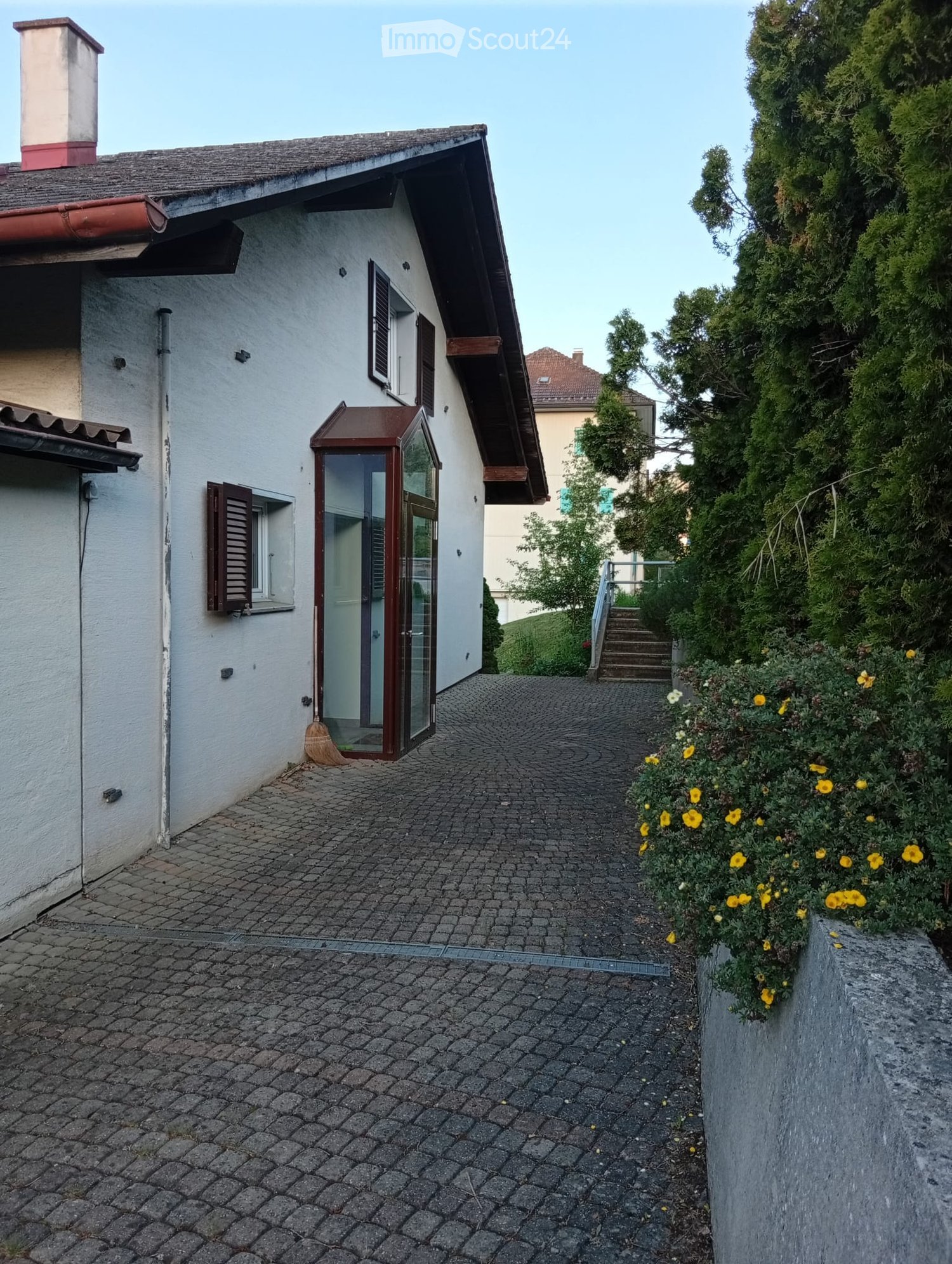 A two-story house with a gray exterior, a red-tiled roof, and a small entrance area with a glass door and wooden shutters. The pathway is paved with cobblestones, and there are yellow flowers in a planter on the right side.