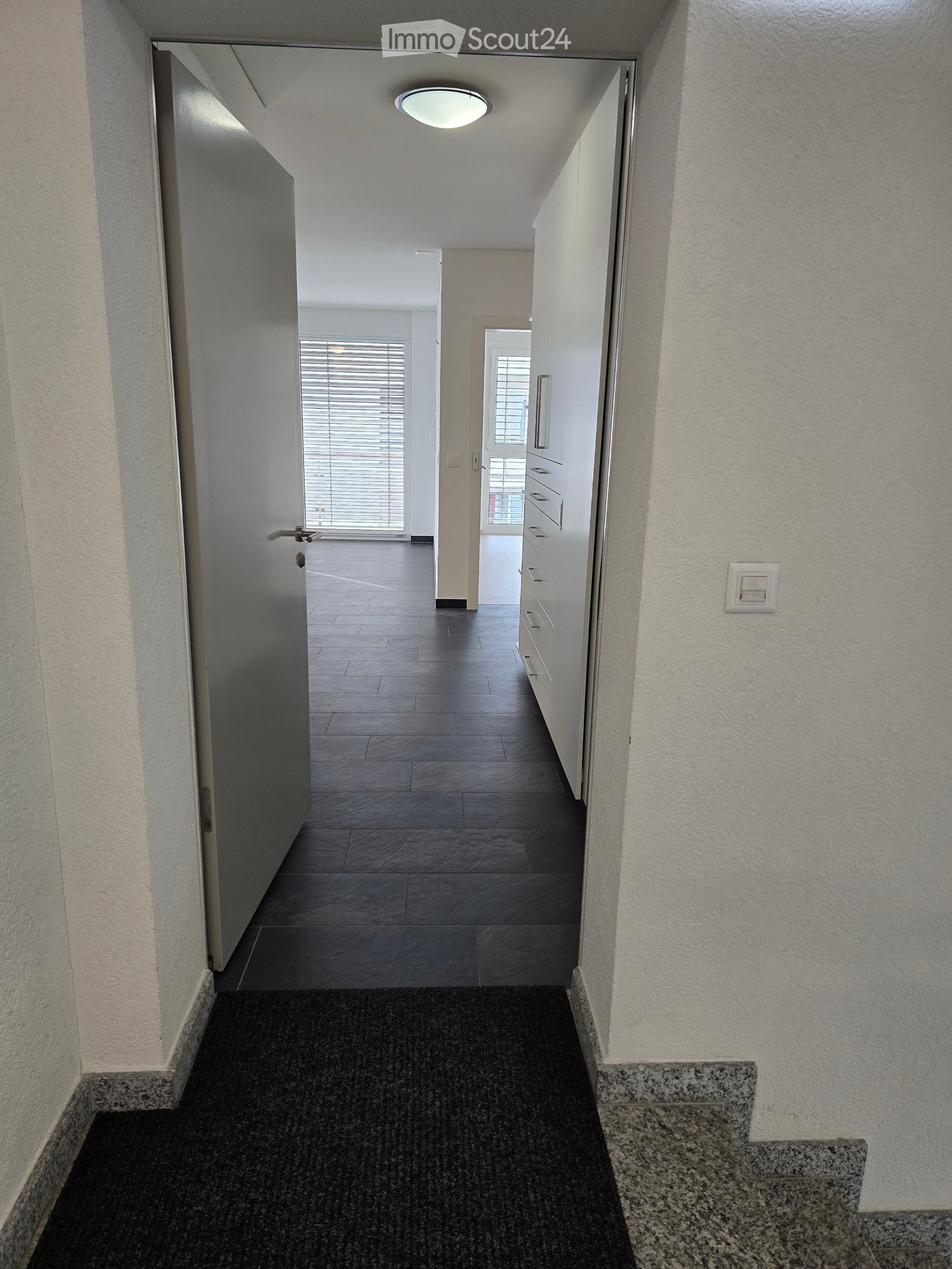 Clean, modern hallway with dark tiled floor, white walls, and a ceiling light. Features a partially open door leading to another room and a staircase with granite steps.