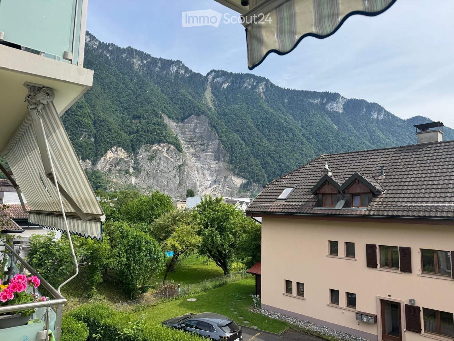 Balcony overlooking a scenic mountain view with a house and garden below.