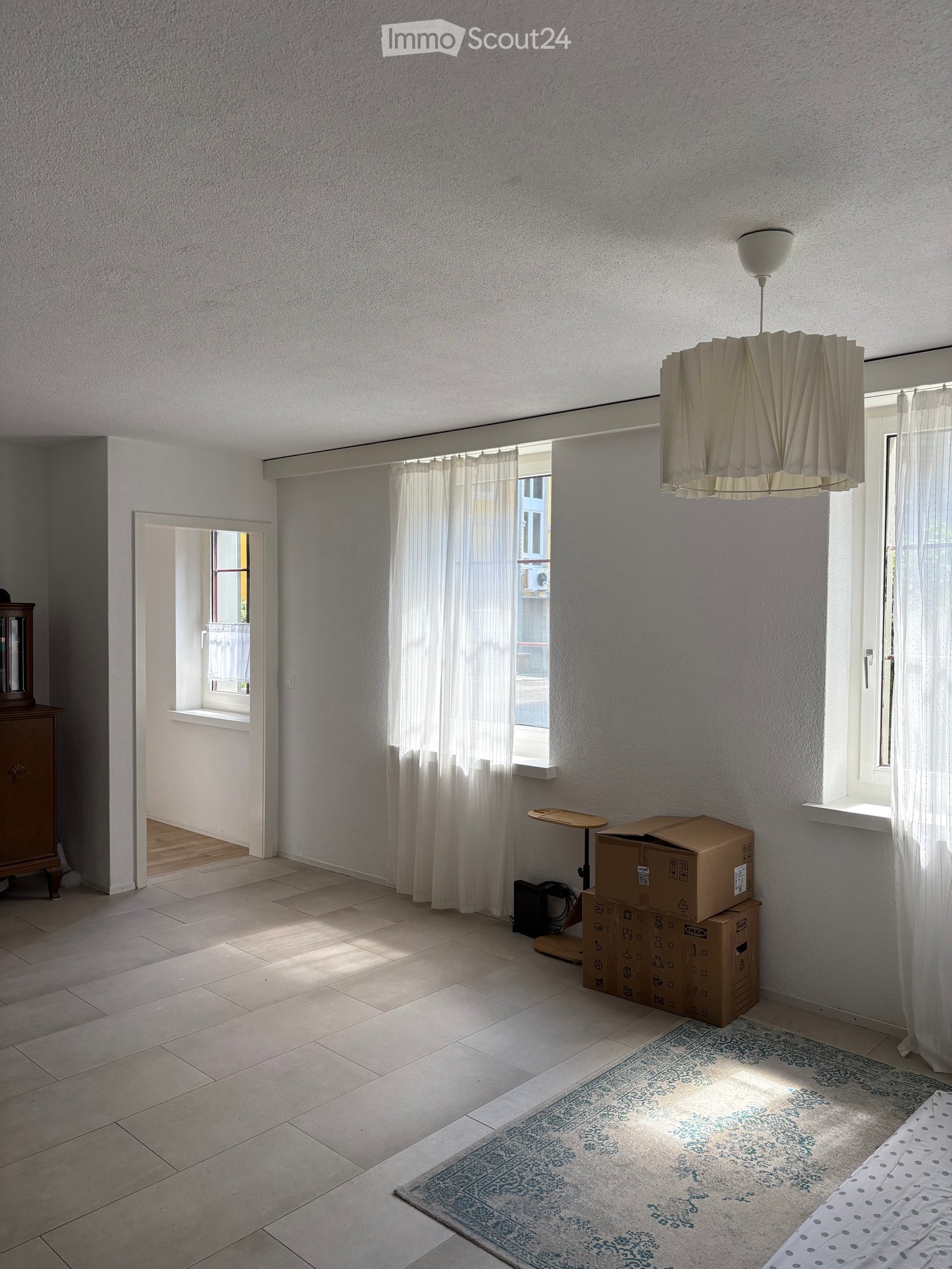 Bright, minimalist room with large windows, pendant light, small table, cabinet, and a bed with a patterned duvet.