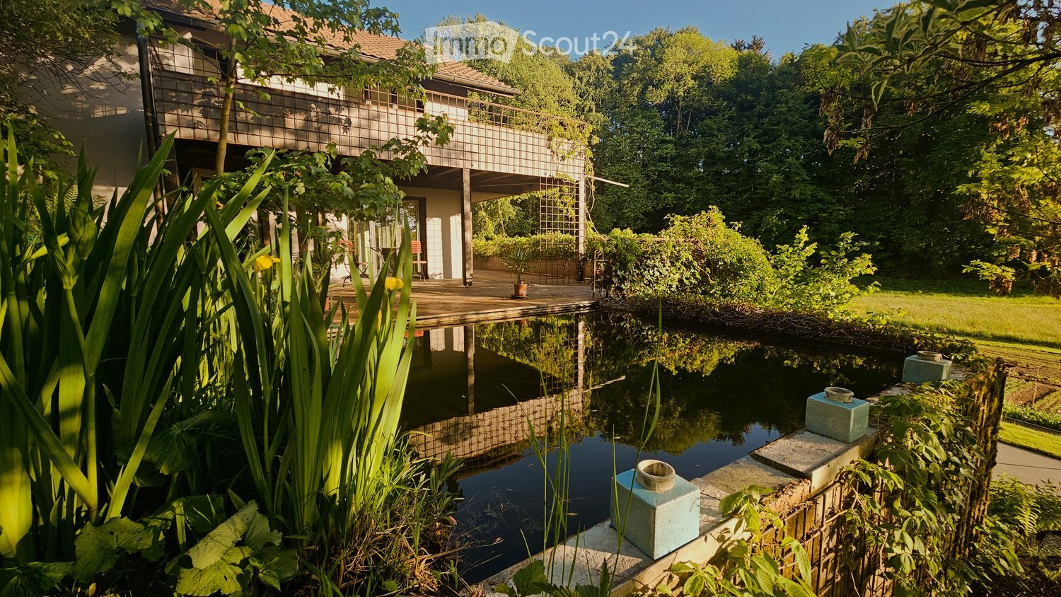 A serene garden with a pond reflecting the modern house, lush greenery, and a wooden deck with outdoor furniture.