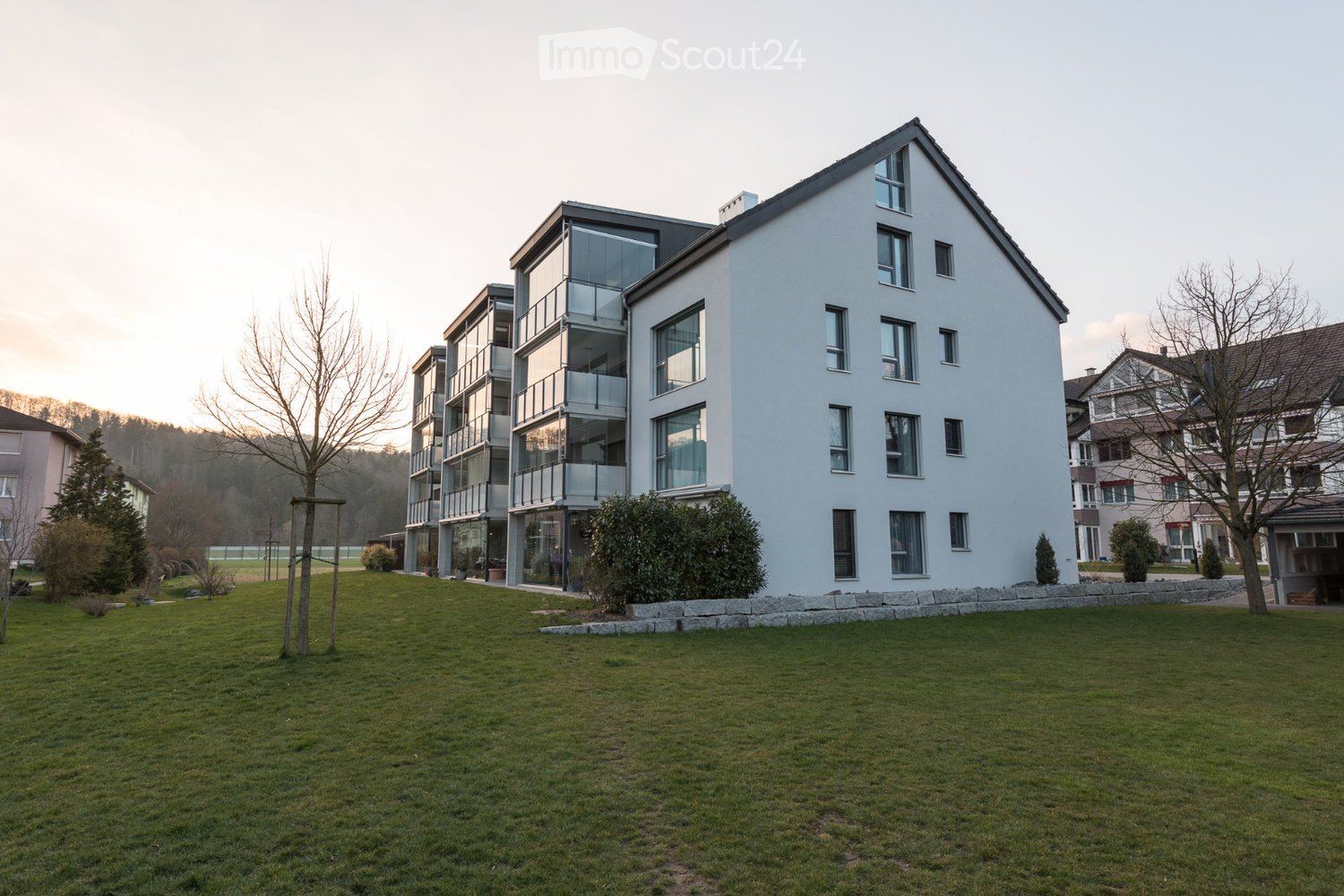 Modern multi-story residential building with white exterior, large windows, and balconies, surrounded by a well-maintained lawn and trees.