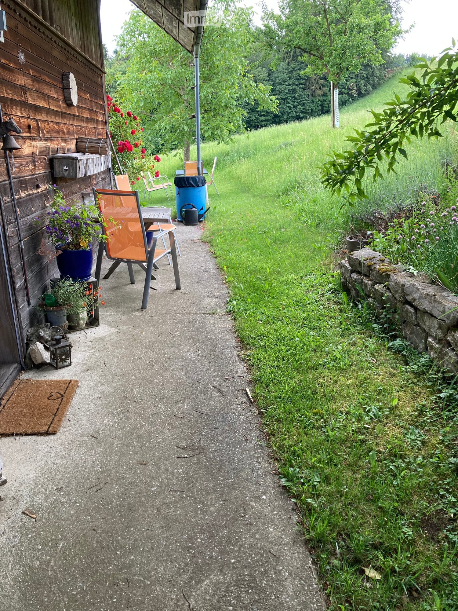 Outdoor pathway leading to a wooden house, with chairs, a table, and potted plants near the entrance.