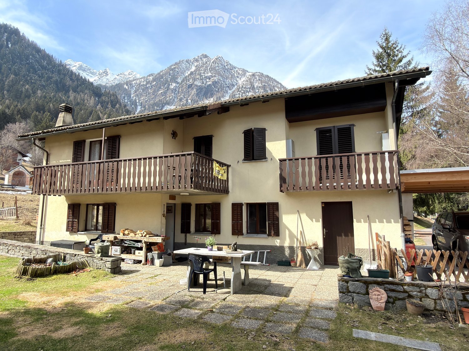 Two-story house with beige walls, wooden shutters, and balconies overlooking a scenic mountain view. The yard includes a table, chairs, and various tools and items.