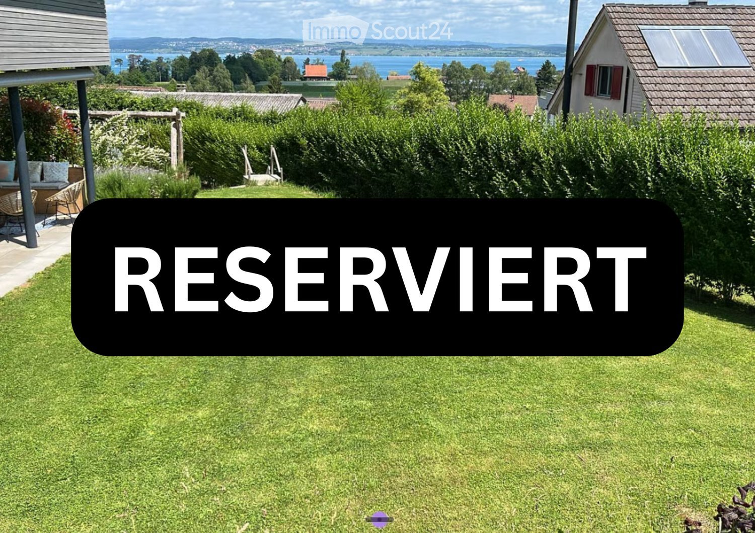 Lush green lawn with a large black sign reading "RESERVIERT" (reserved) in the center, surrounded by trees, shrubs, and a view of a lake and distant mountains.