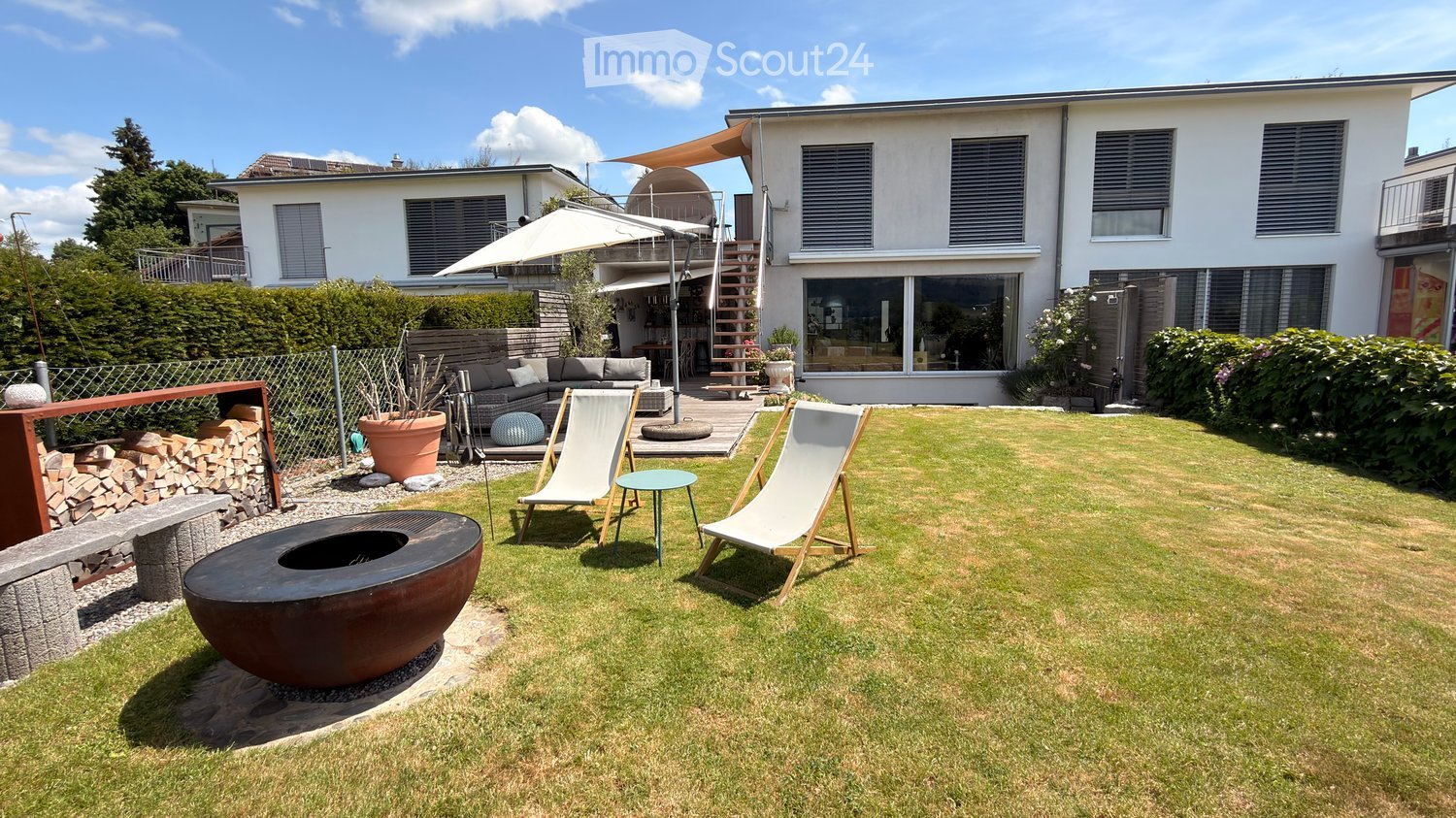 Well-maintained garden with lawn, fire pit, lounge chairs, small table, and a modern house in the background.