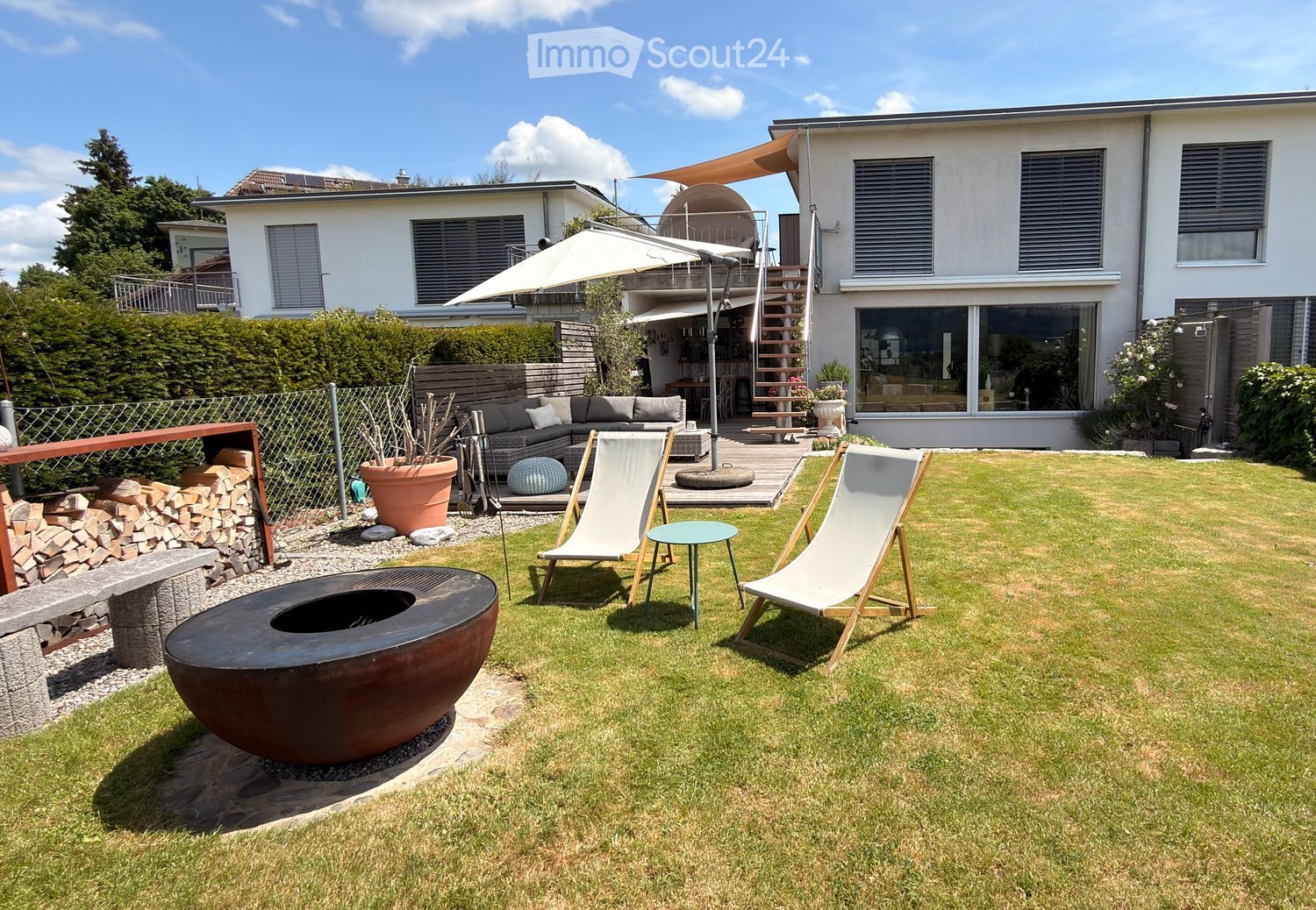 Well-maintained garden with a fire pit, lounge chairs, a small table, a sofa, and a large umbrella. The area is enclosed by a fence and features a modern house in the background.