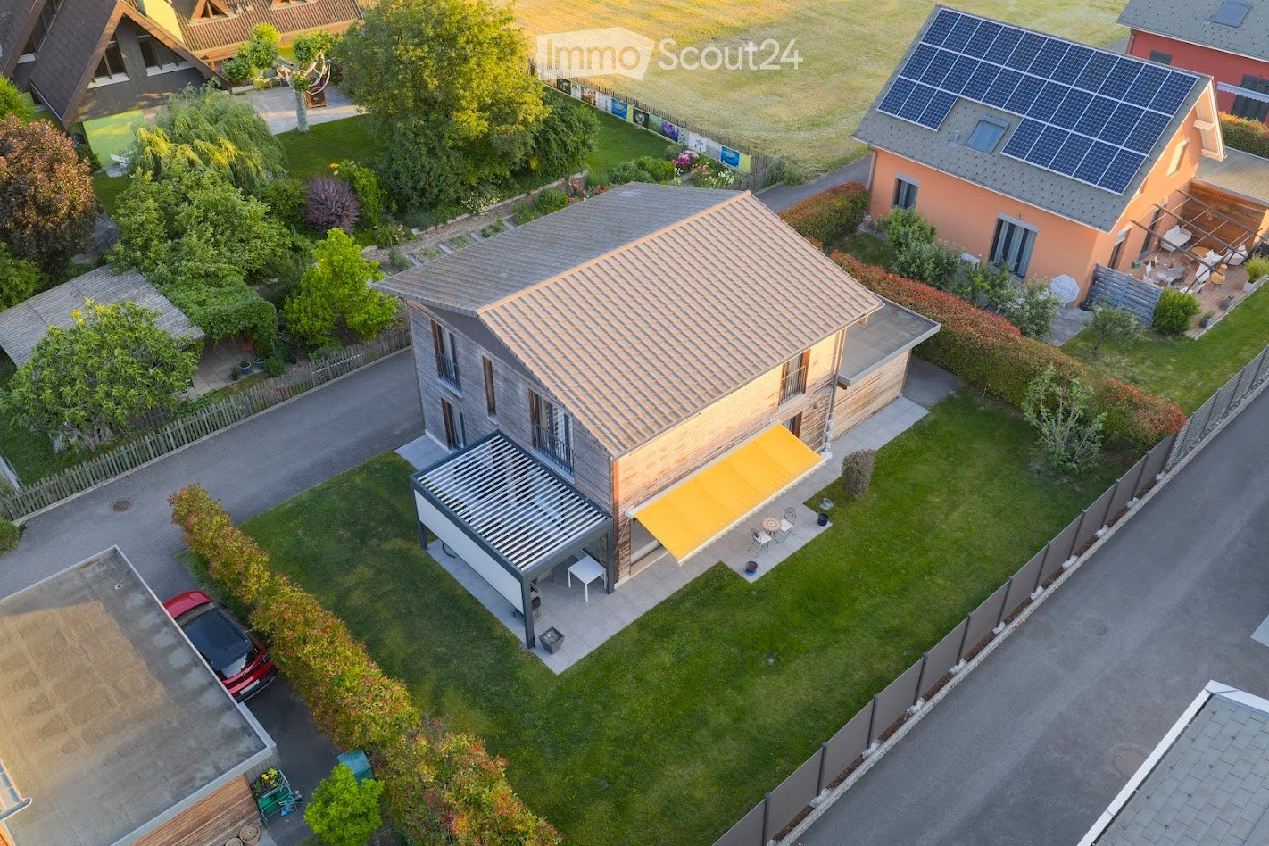Aerial view of a modern two-story house with a grey tiled roof, surrounded by a well-maintained garden, a covered patio with outdoor furniture, and solar panels on the roof of a neighboring house.