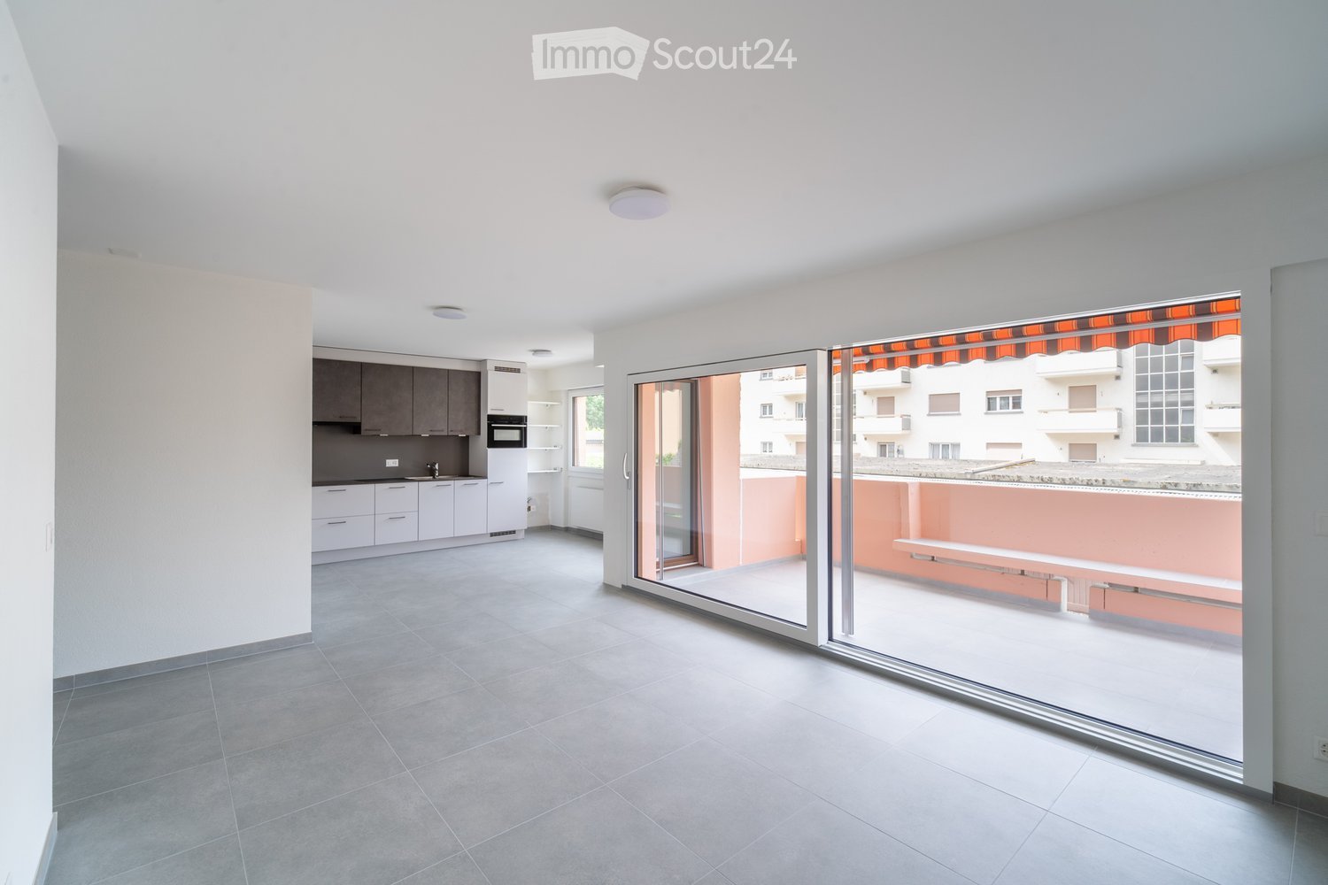 Modern, unfurnished living space with a fully equipped kitchen area, large sliding glass doors leading to a balcony, and grey tiled flooring.