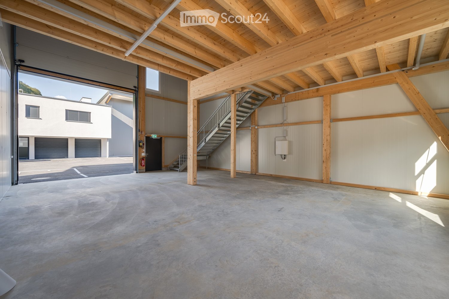 Empty warehouse space with exposed wooden beams, concrete floor, and a staircase leading to an upper level. The space is well-lit with natural light from large windows and has a modern, industrial design.