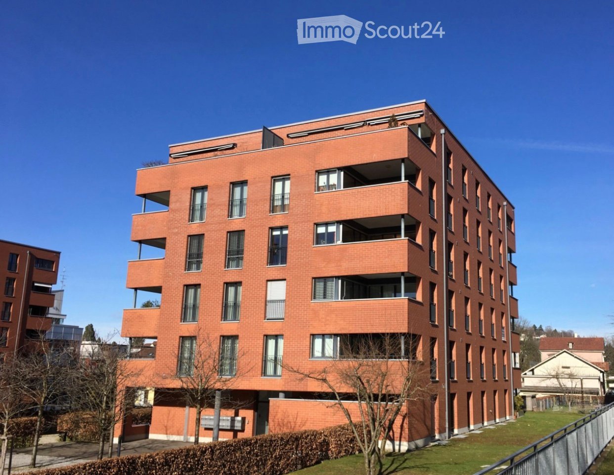 Modern multi-story apartment building with red brick facade, multiple balconies, and large windows.