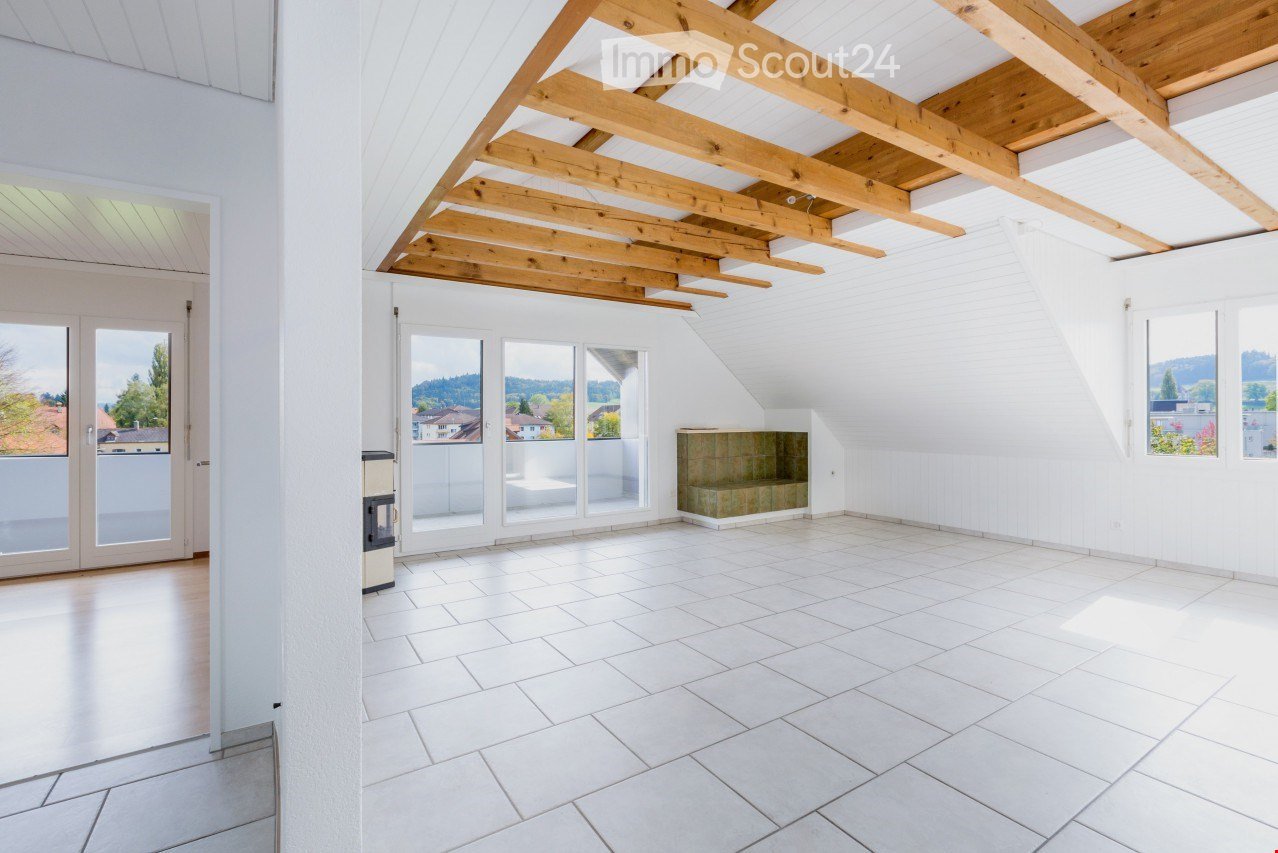 Bright, empty attic room with white walls, wooden beams, large windows, and a tiled floor. No furniture or equipment present.