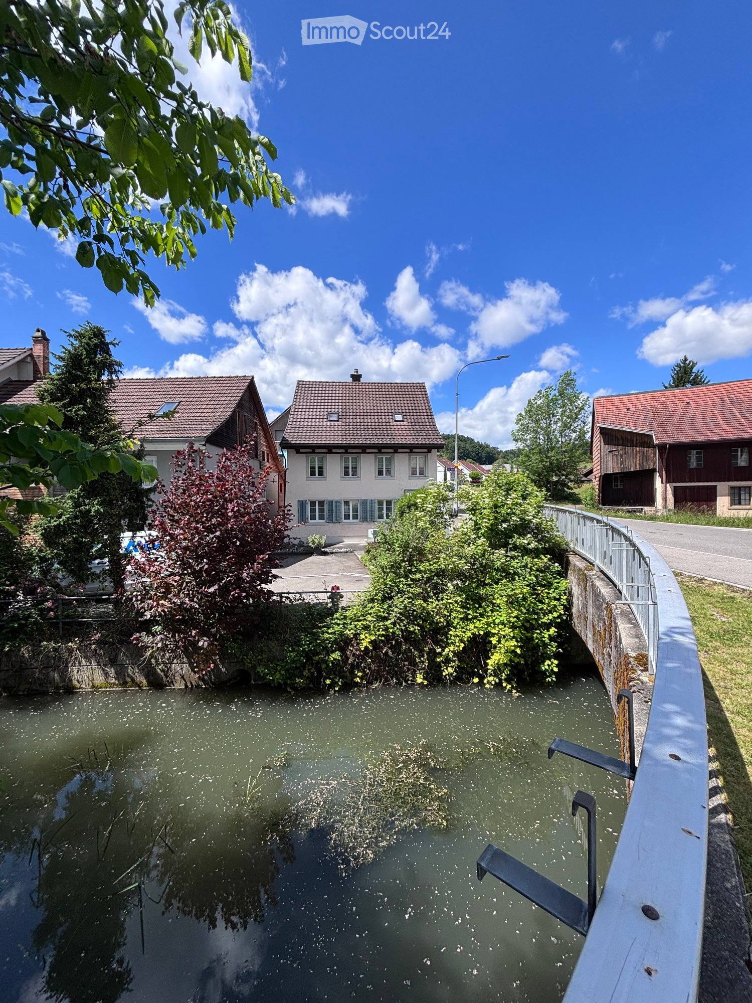 Residential area with houses, a canal, and greenery. Houses have red-tiled roofs and white walls, surrounded by trees and shrubs.