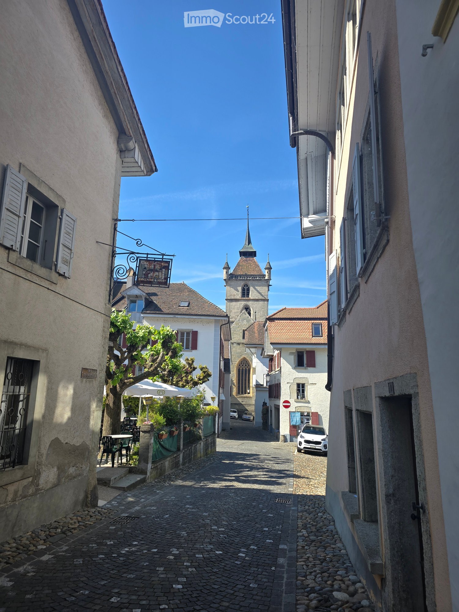 A narrow cobblestone street lined with old buildings, featuring a church tower in the background and a few parked cars.