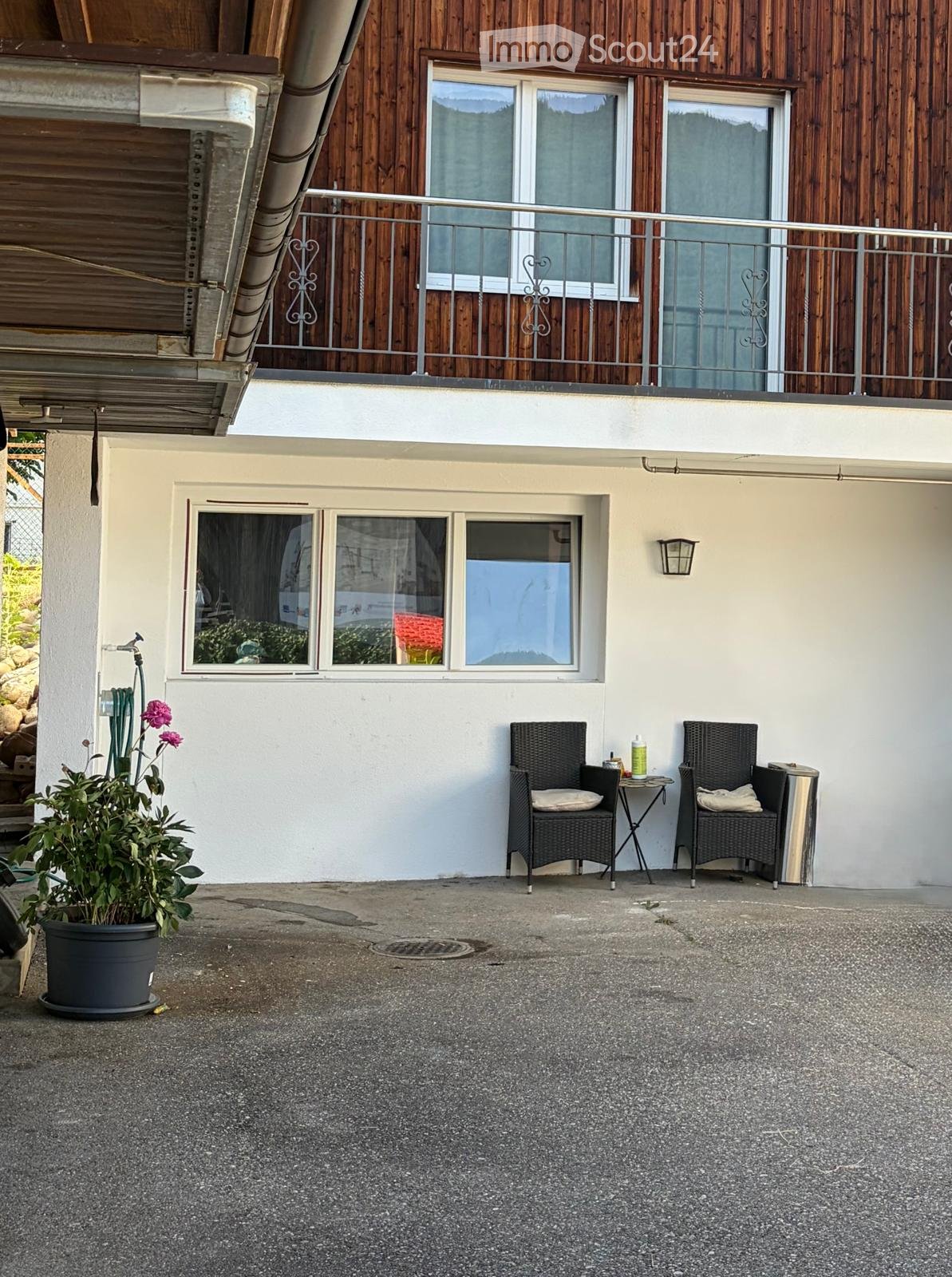 Balcony with two wicker chairs, a small table, and a potted plant. The balcony is attached to a building with wooden siding and large windows.
