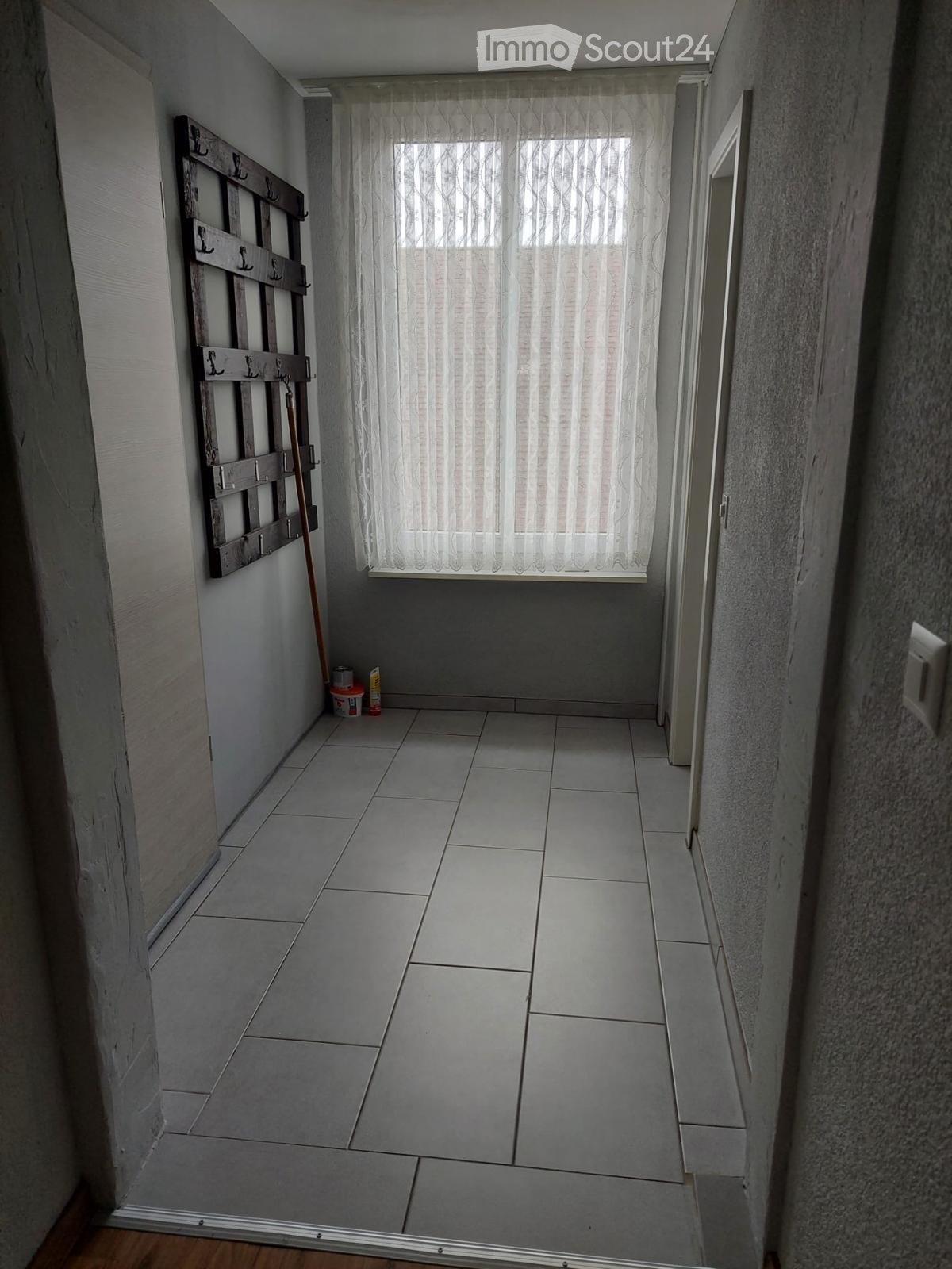 Empty hallway with tiled floor, white walls, and a window with sheer curtains. A coat rack and cleaning supplies are visible on the left side.