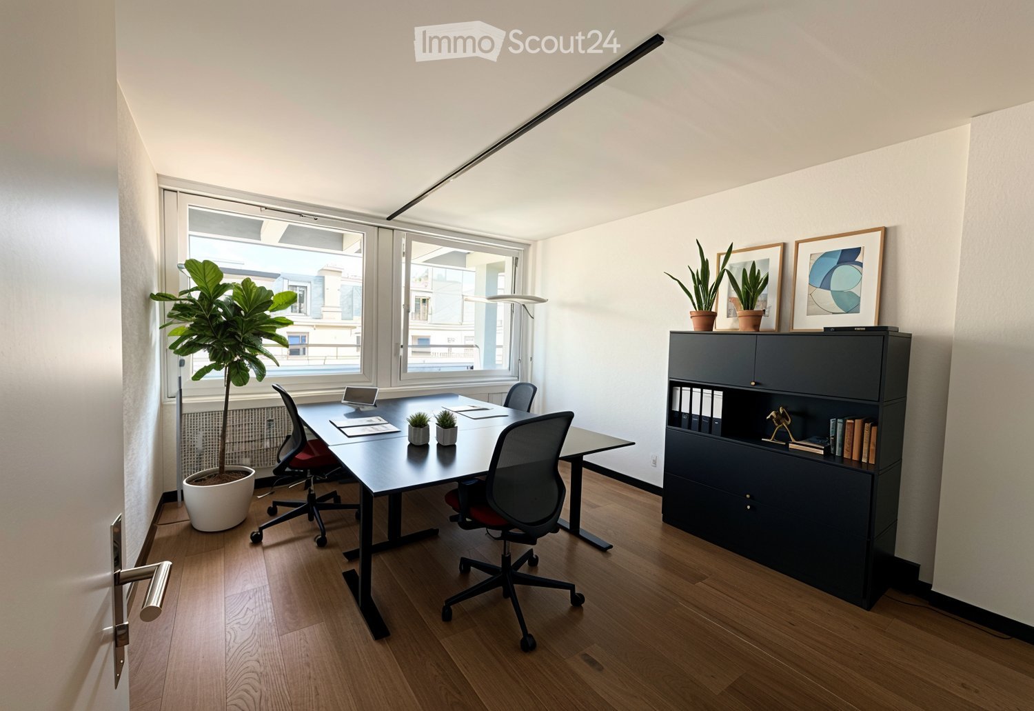Modern office space with a large wooden desk, ergonomic chairs, a dark cabinet, potted plants, and framed artwork on the wall. Large windows provide natural light.