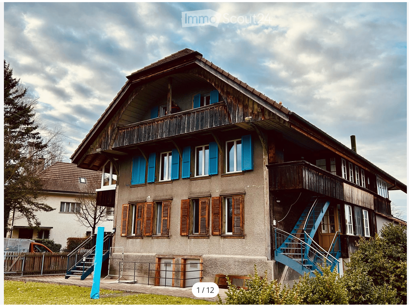 Two-story house with wooden balconies, blue shutters, and a staircase leading to the entrance.