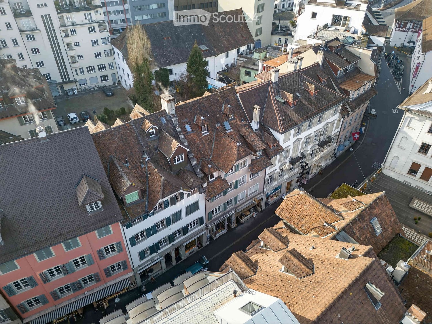 Aerial view of a dense urban area with multiple buildings featuring traditional red-tiled roofs, chimneys, and chimneys, surrounded by narrow streets and parked cars.