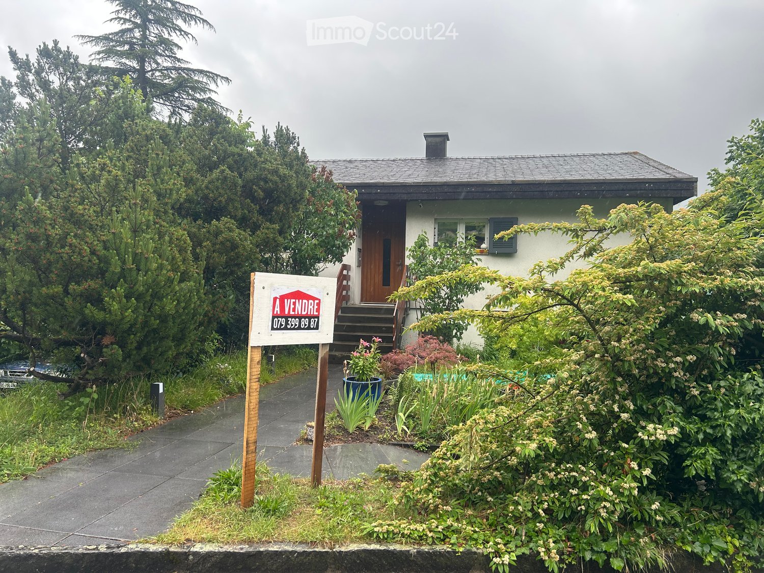 A two-story house with a grey roof, white walls, and a wooden front door, surrounded by lush greenery and a "A VENDRE" sign indicating it is for sale.
