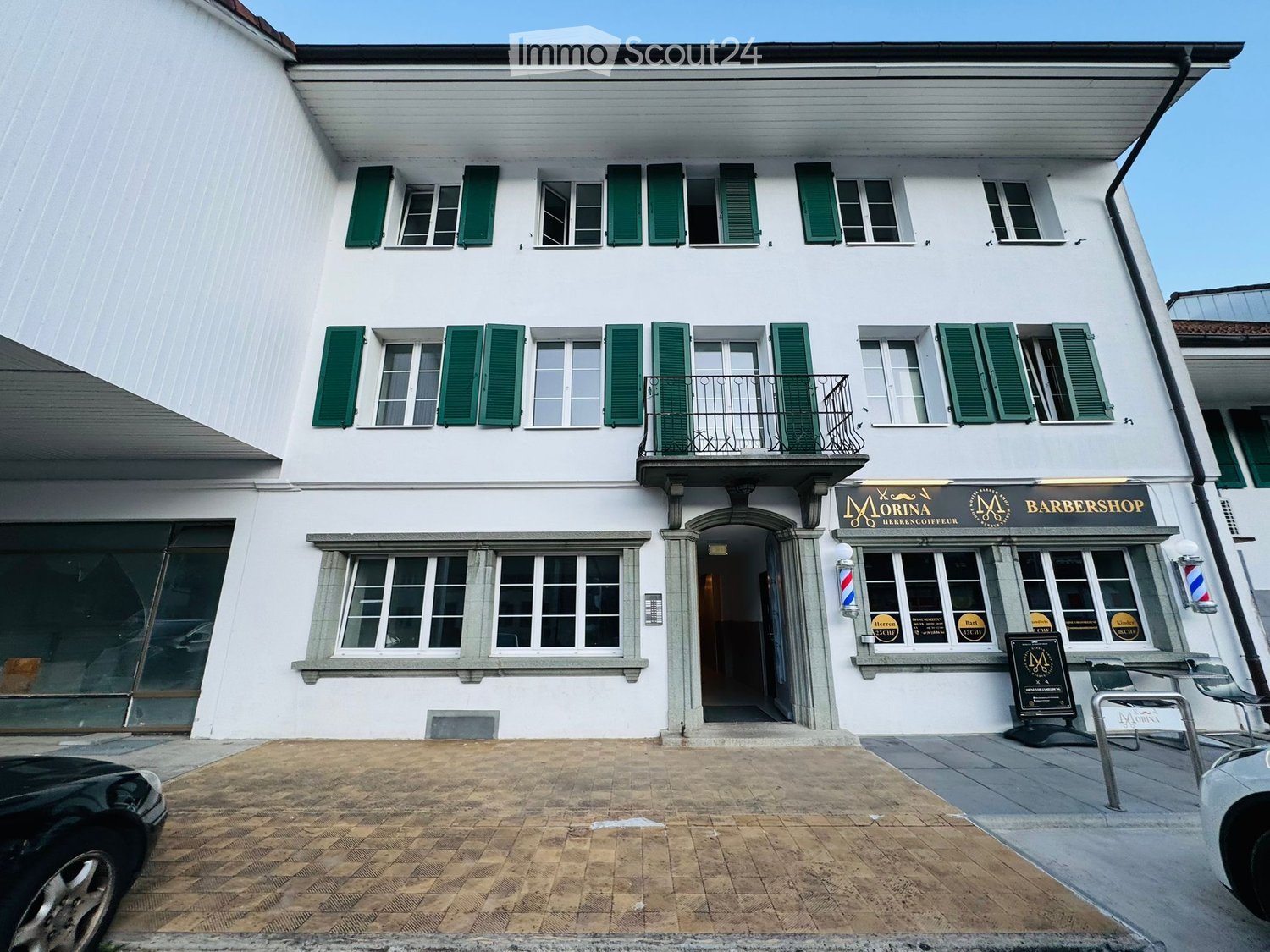 A two-story white building with green shutters, featuring a balcony on the second floor and a storefront labeled 'Morina Barbershop' on the ground floor.