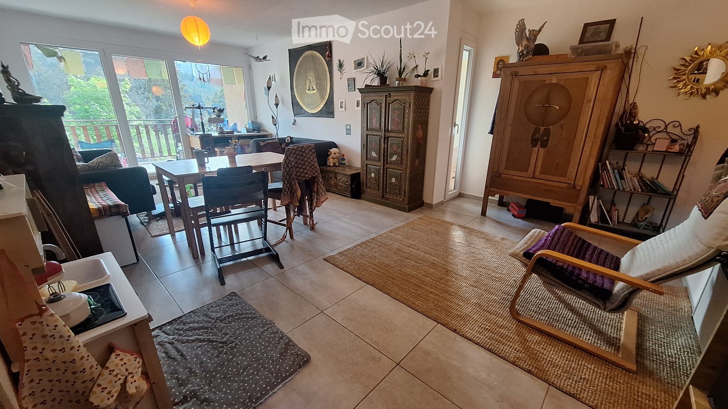 Bright living room with large windows and a balcony view, featuring a sofa, chairs, a dining table, a cabinet, shelves, and decorative items.