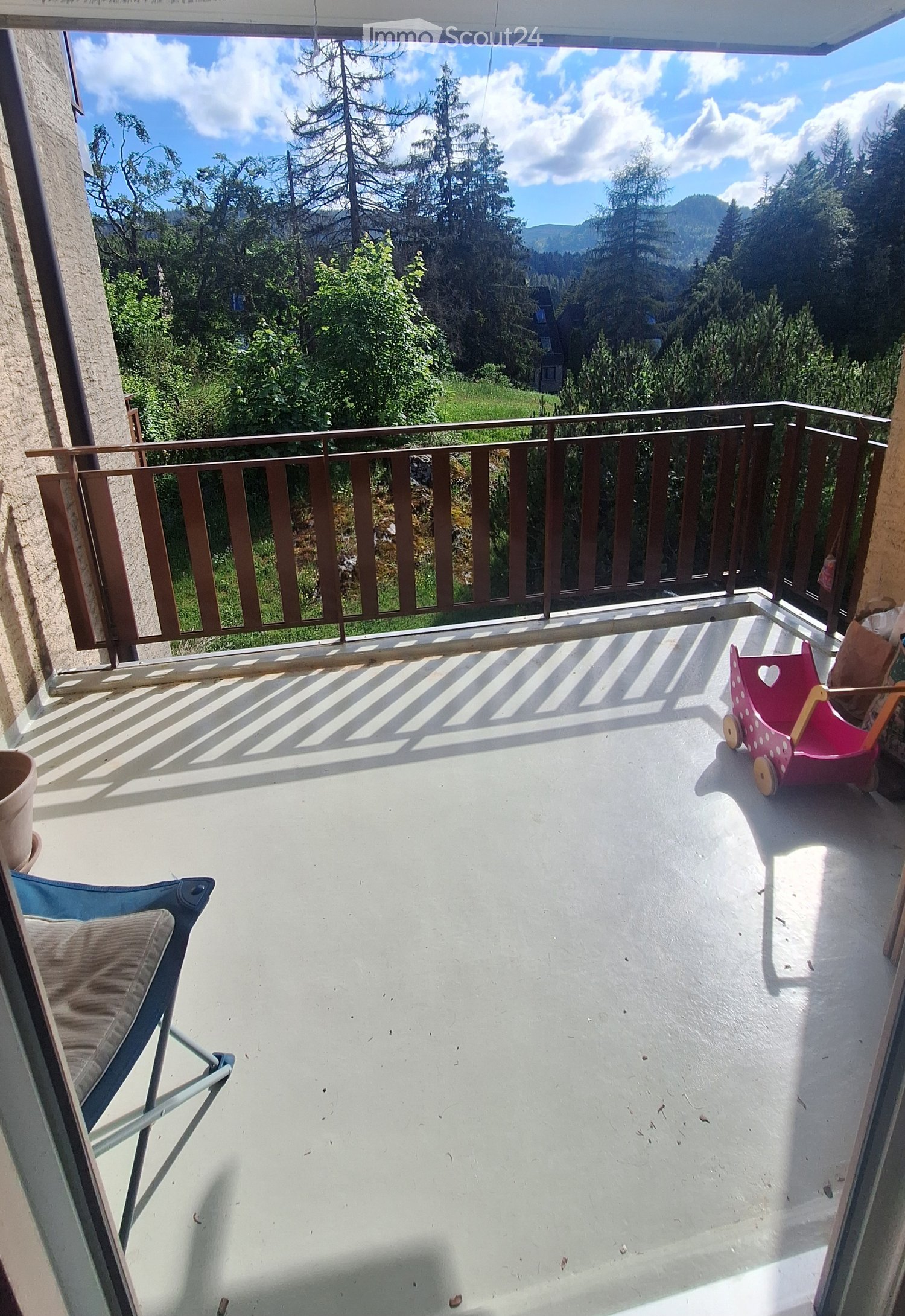 Balcony with a scenic mountain view, featuring a metal railing, a blue folding chair, and a pink toy cart.