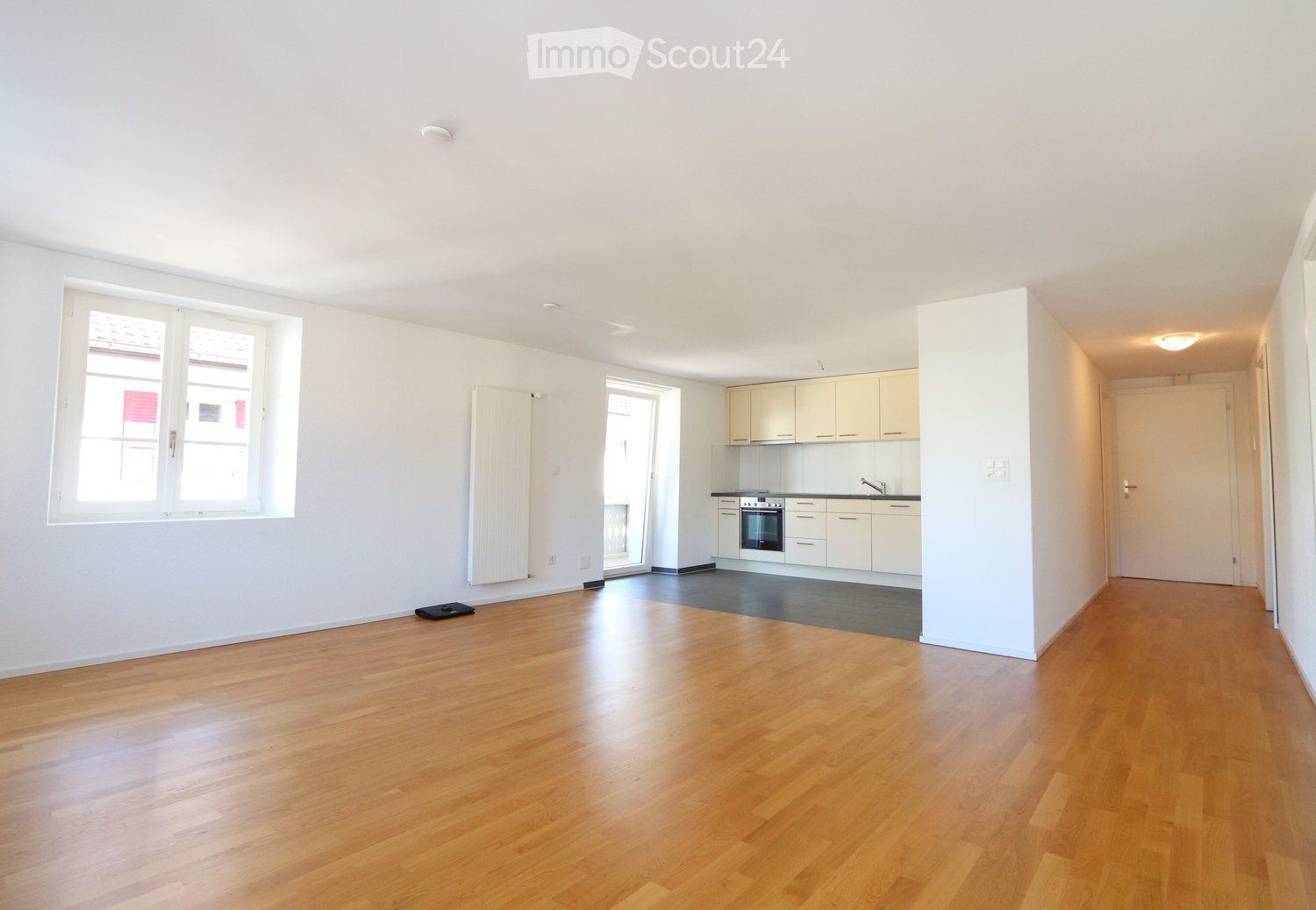 Bright, unfurnished room with wooden flooring, white walls, and a small kitchen area featuring cabinets, an oven, and a sink.