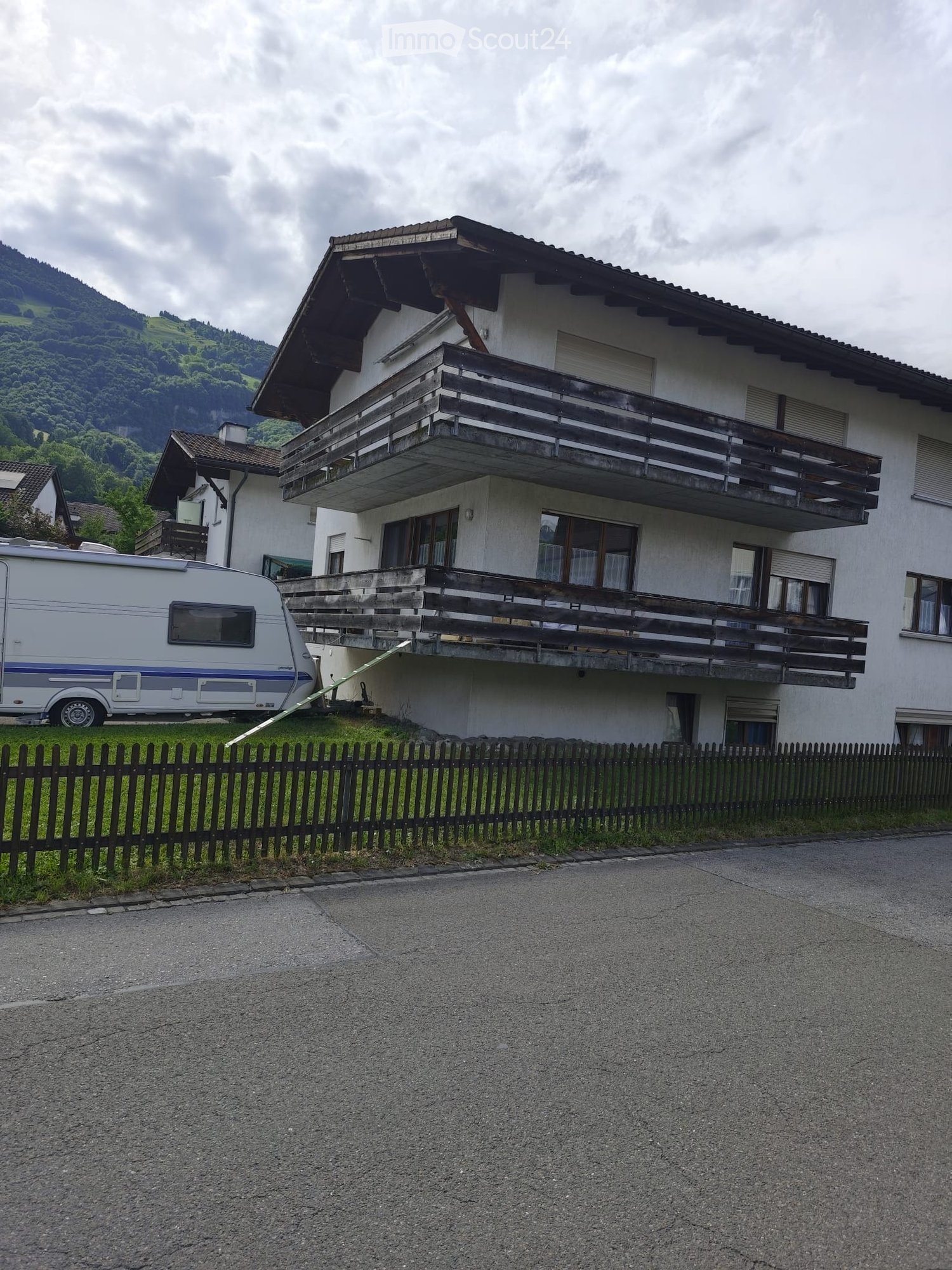 Two-story house with a balcony, surrounded by a wooden fence and a trailer parked nearby.