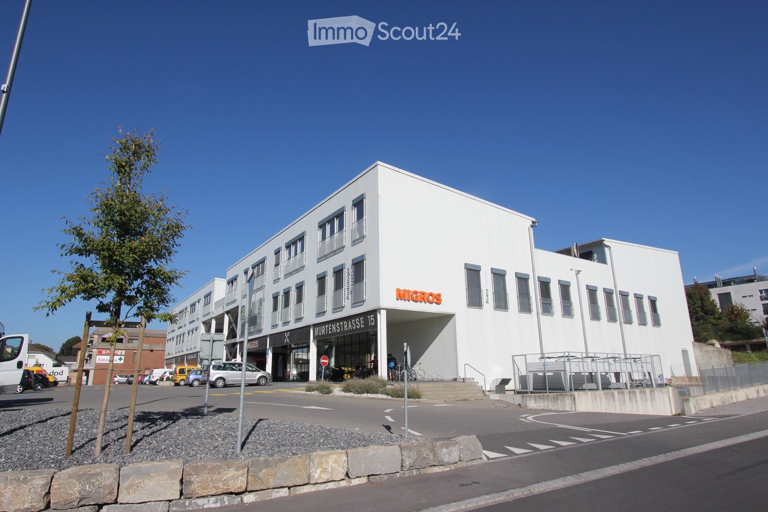 a large white building with several windows, a sign that reads 'Migros', a car parked in front of the building, a staircase leading to the entrance, and some street lights