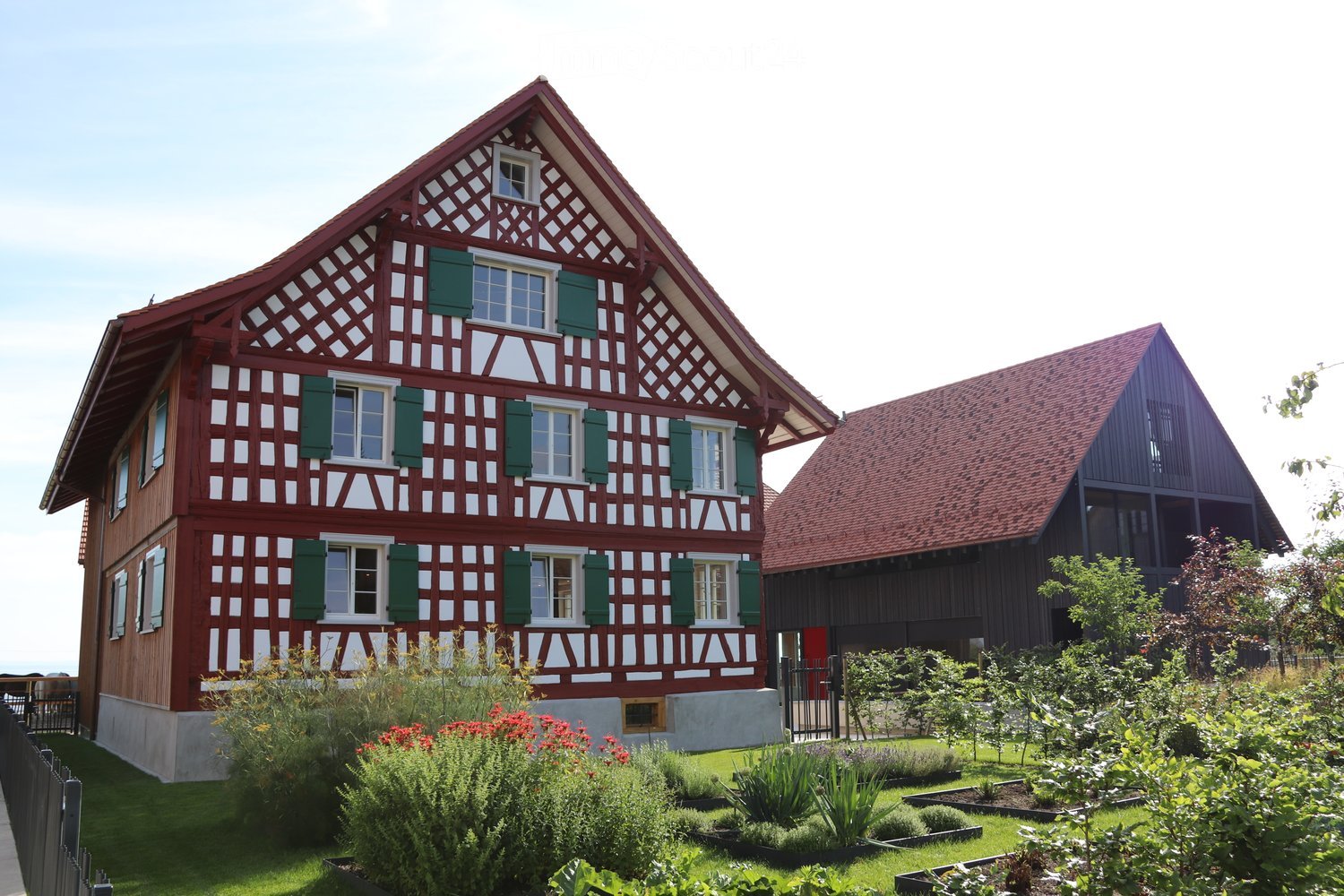 Traditional half-timbered house with red and white exterior, green shutters, and a well-maintained garden with various plants and shrubs.