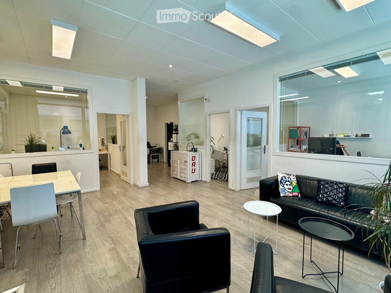 Modern, open-plan office space with light wood flooring, white walls, and large windows. Features a black leather sofa with colorful cushions, a small round white side table, a black armchair, a small black coffee table, a white dining table with chairs,
